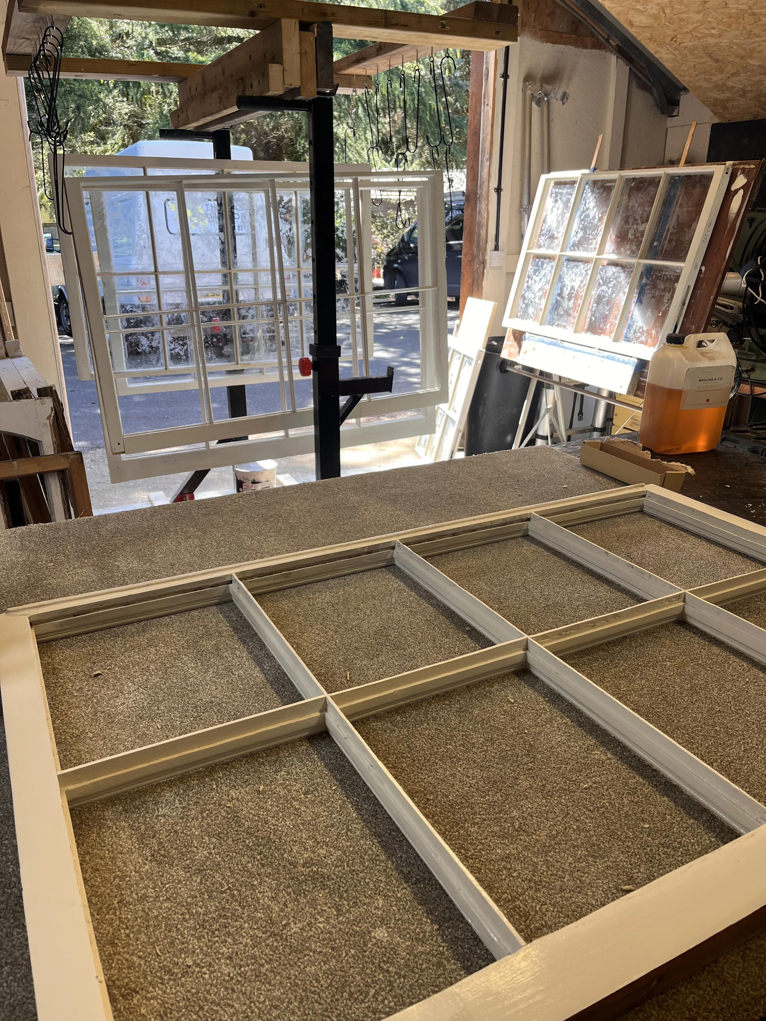 Workshop scene with white window frames being assembled or repaired. In the background, there are multiple large glass window panes leaning against a wooden wall, and a sawhorse supports one of the window frames. The workspace is outdoors with a view of trees and parked cars visible through the open garage door.