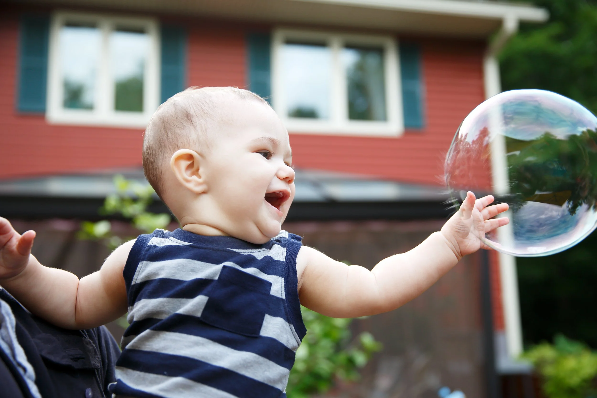 Baby boy in striped blue tank top reaching for a bubble in front of a red house with green shutters
