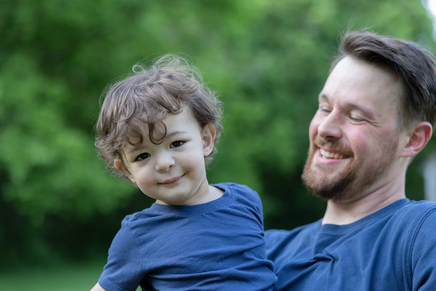 Father holding smiling at son with green grass and trees in background