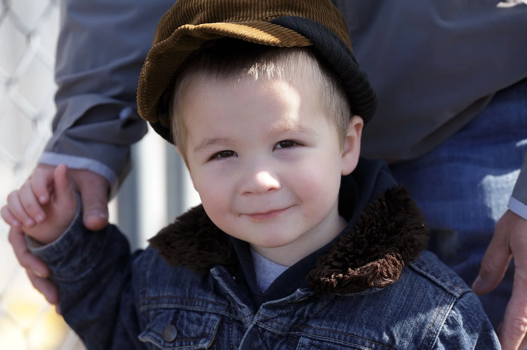 Small boy in denim jacket and brown hat looking at the camera with an adult holding his hands