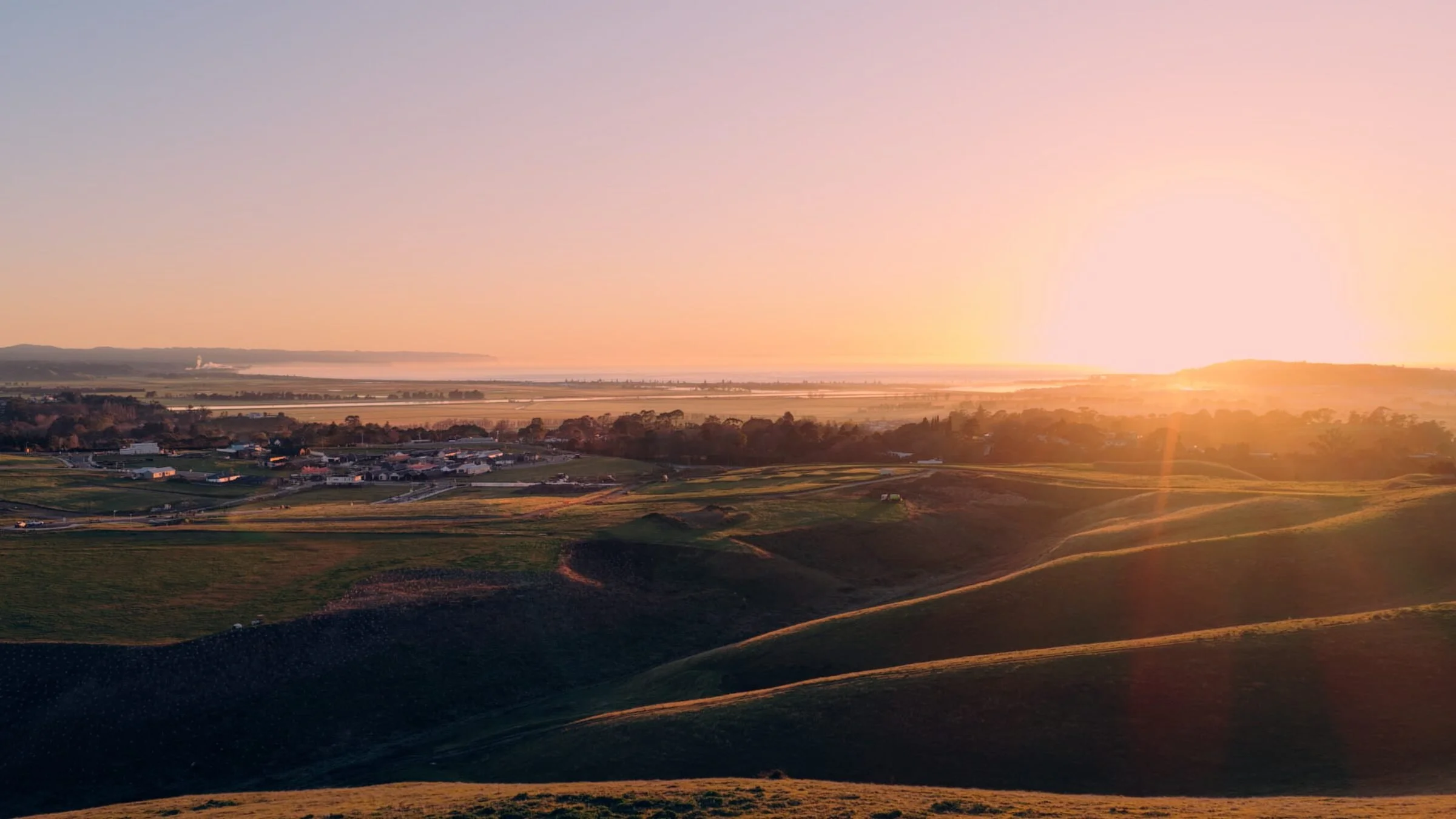 A scenic landscape during sunset with rolling green hills, a small settlement, and a body of water in the distance, all illuminated by warm sunlight.
