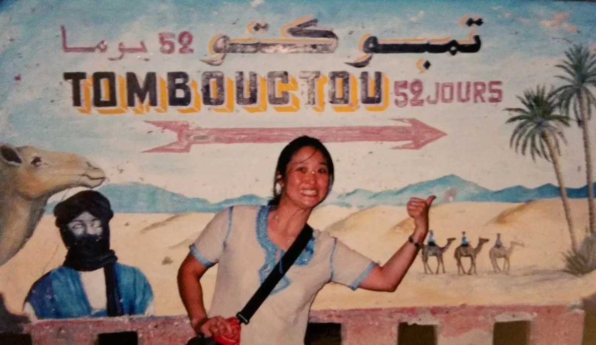 Linda smiling and pointing toward the “52 days to Timbuktu” sign in Zagora, Morocco, painted with a colorful Sahara desert scene of camels, palm trees and a camel herder.