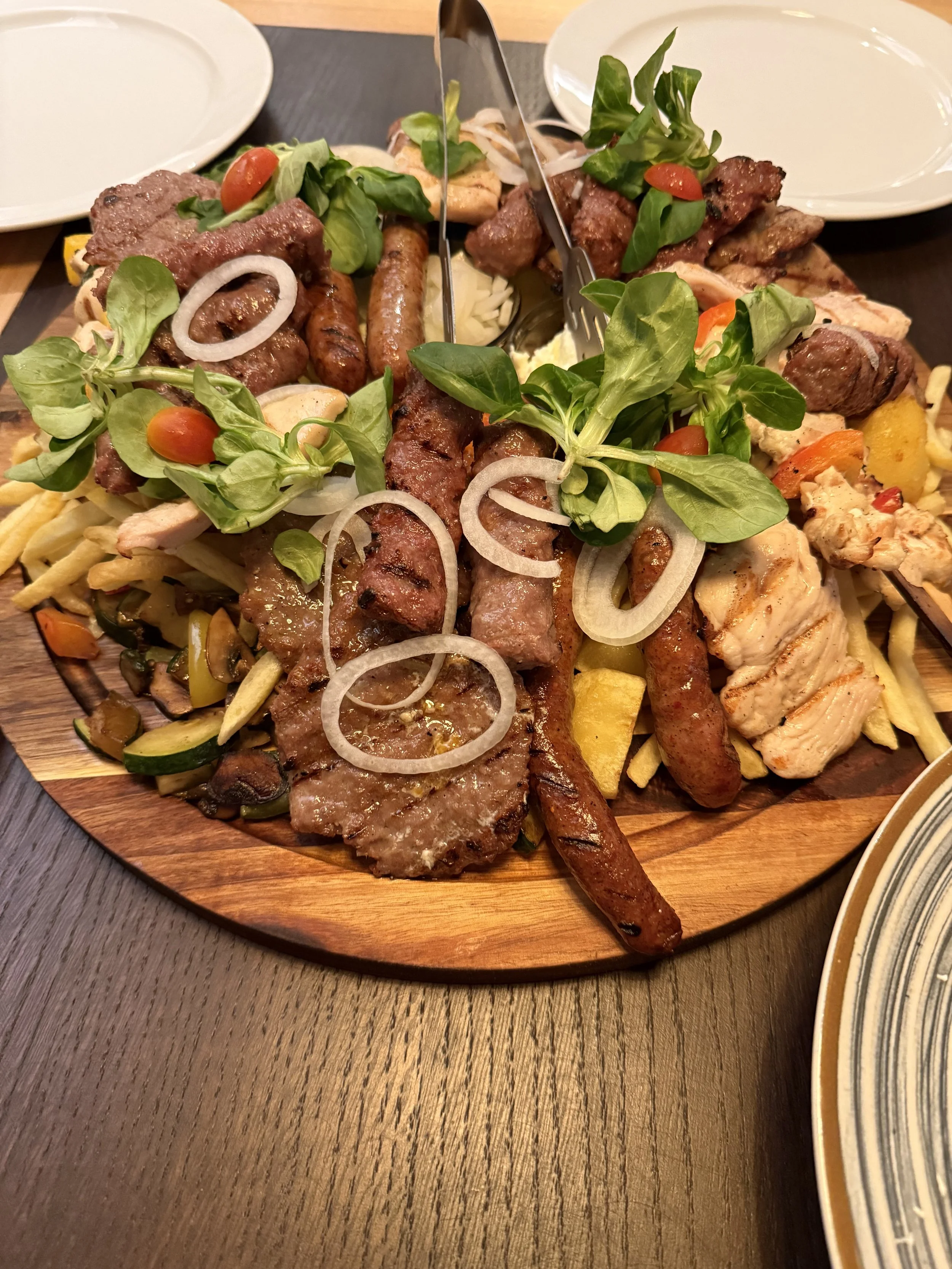 Ein runder Holzteller mit gegrilltem Fleisch, Würstchen, frischen Kräutern, Tomaten, Zwiebeln und Paprika. Im Hintergrund sind leere weiße Teller und ein dunkler Tisch sichtbar.