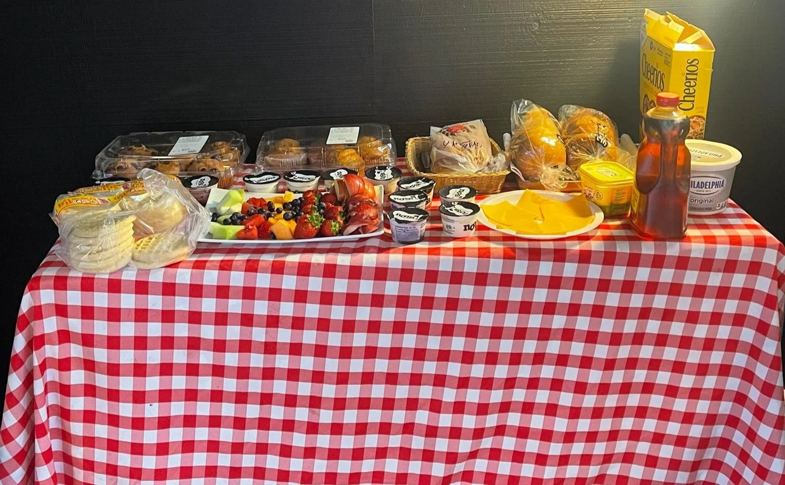 A table covered with a red and white checkered tablecloth holding a variety of breakfast foods including strawberries, blueberries, fruit salad, containers of yogurt, and packaged muffins, along with bread, cheese, butter, orange juice, and cereal.