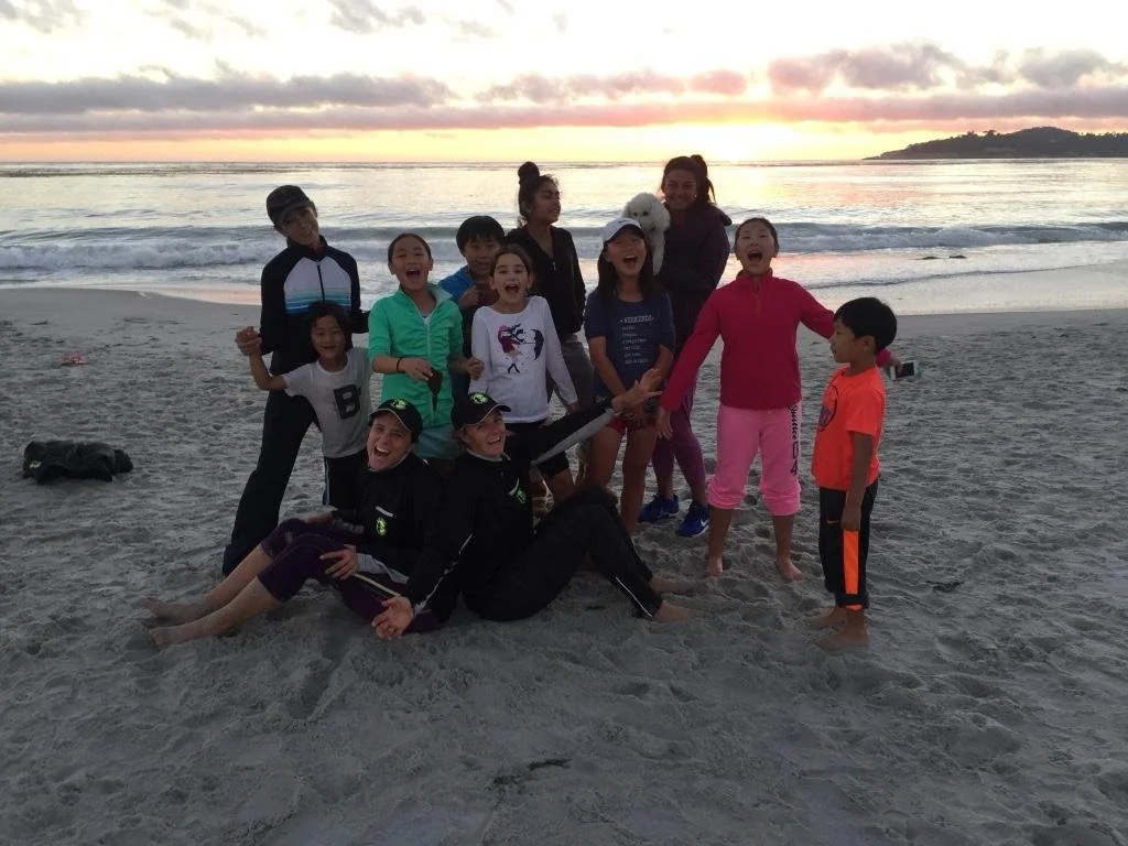 Group of children and two adults on the beach at sunset, smiling and playing near the ocean.