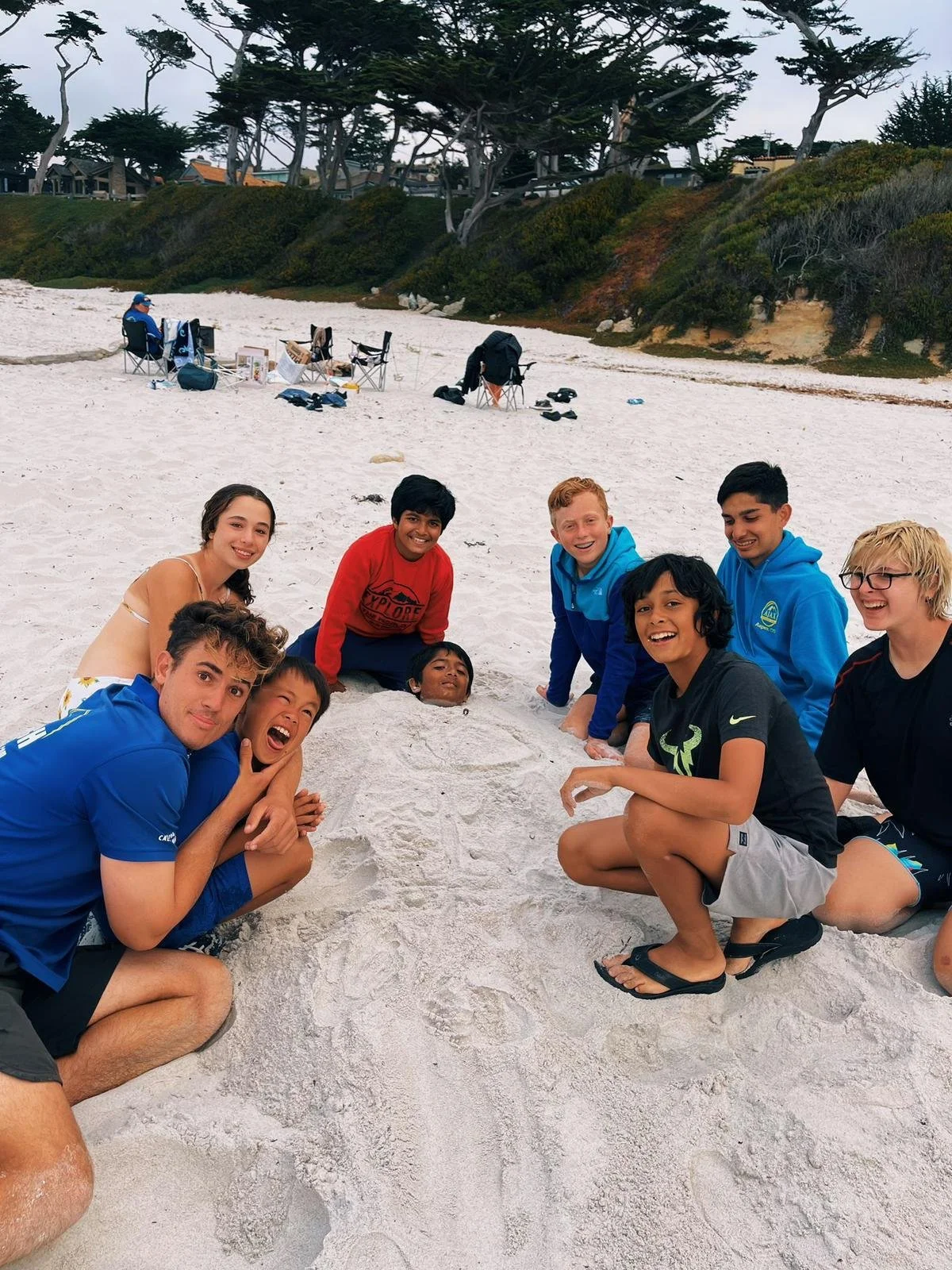 Children and teenagers in a circle on the beach, digging with their hands in the sand and smiling, with beach chairs and trees in the background.