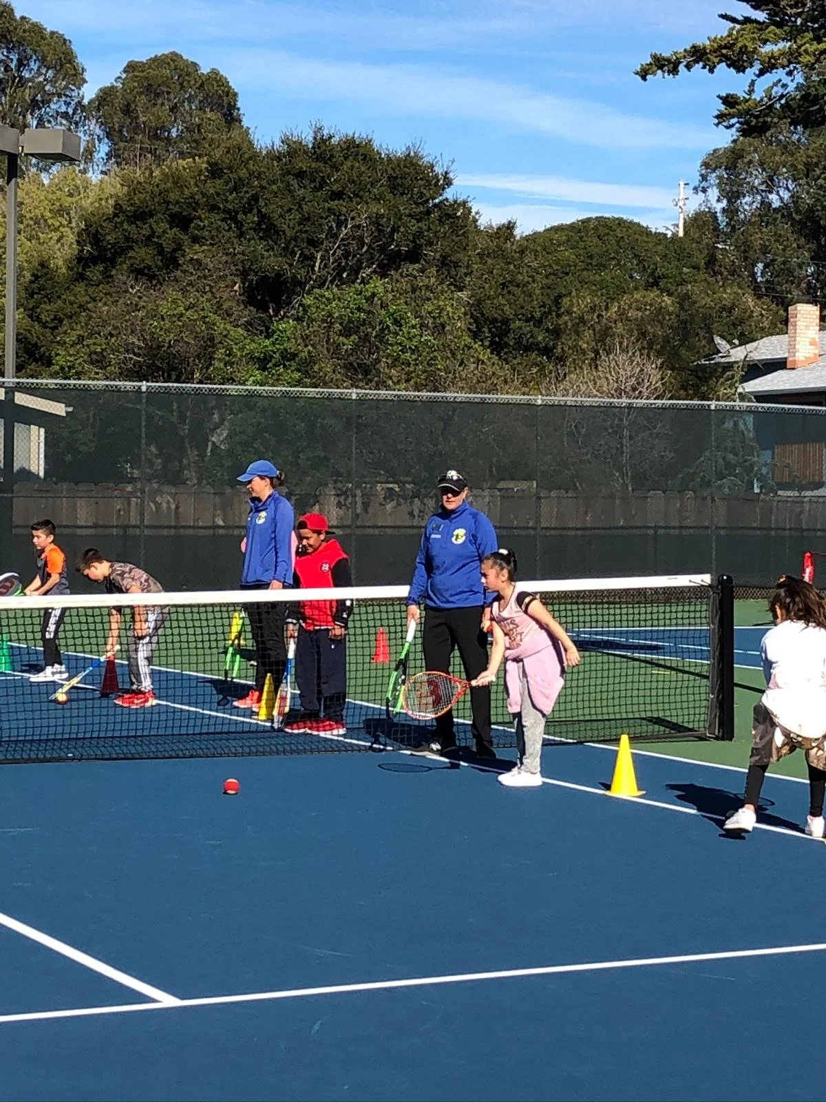 Children and coaches on a tennis court participating in a tennis lesson, with cones and tennis balls present.