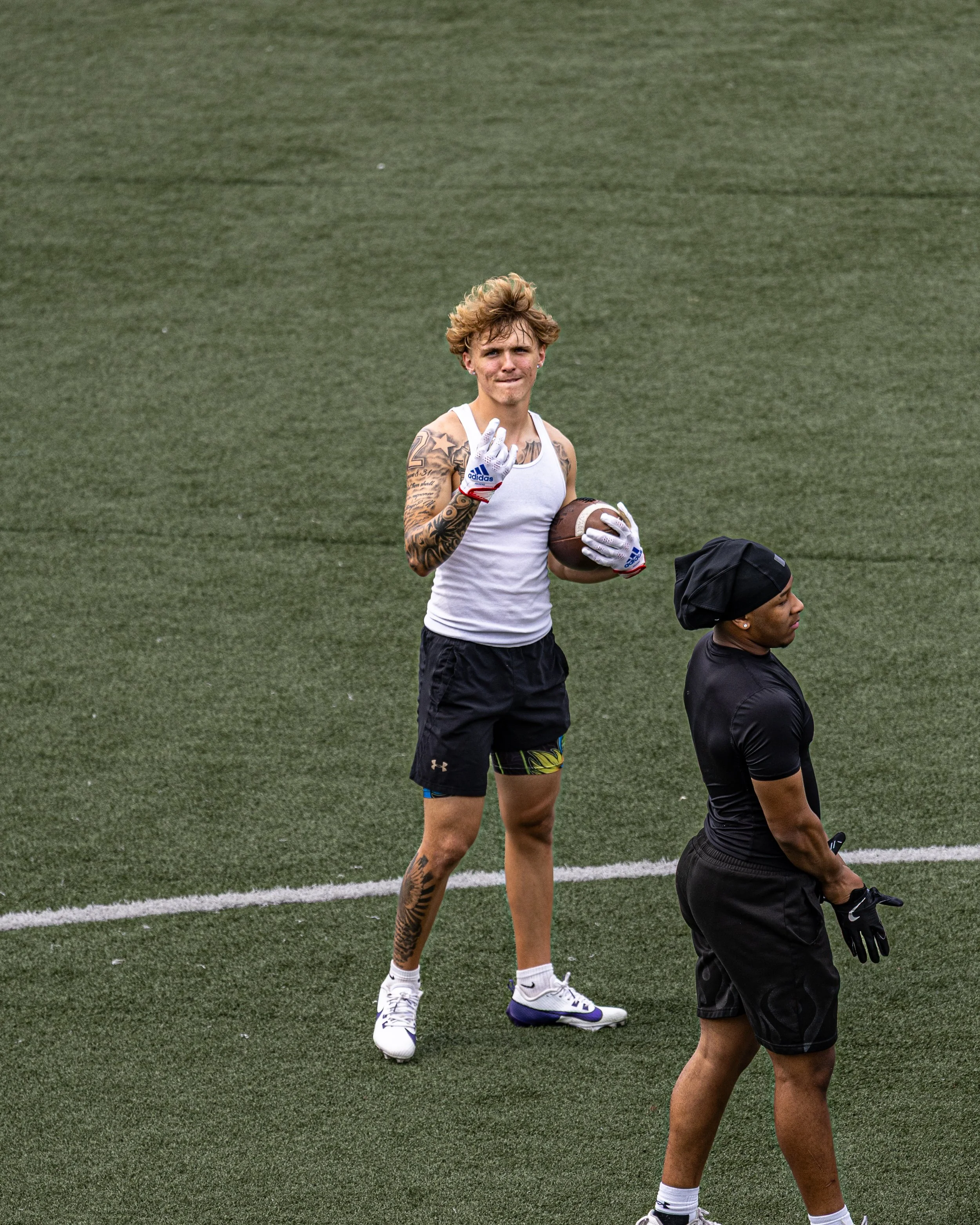 A young man with tattoos holding a football on a green field, making a peace sign, with another woman in black attire and head covering nearby.