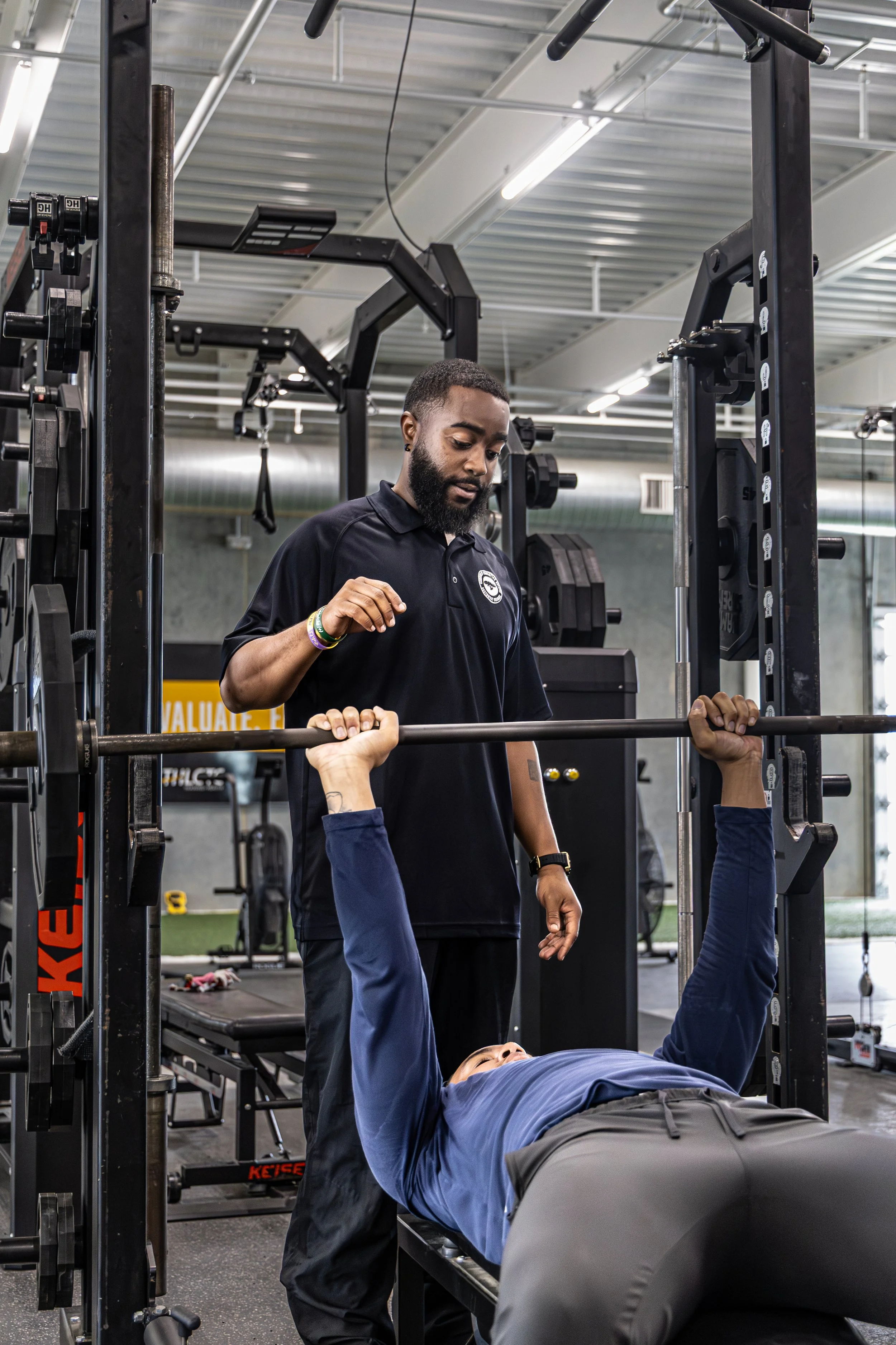 A personal trainer assisting a person with a bench press in a gym. The trainer is standing beside the person lying on a bench, helping with the exercise.