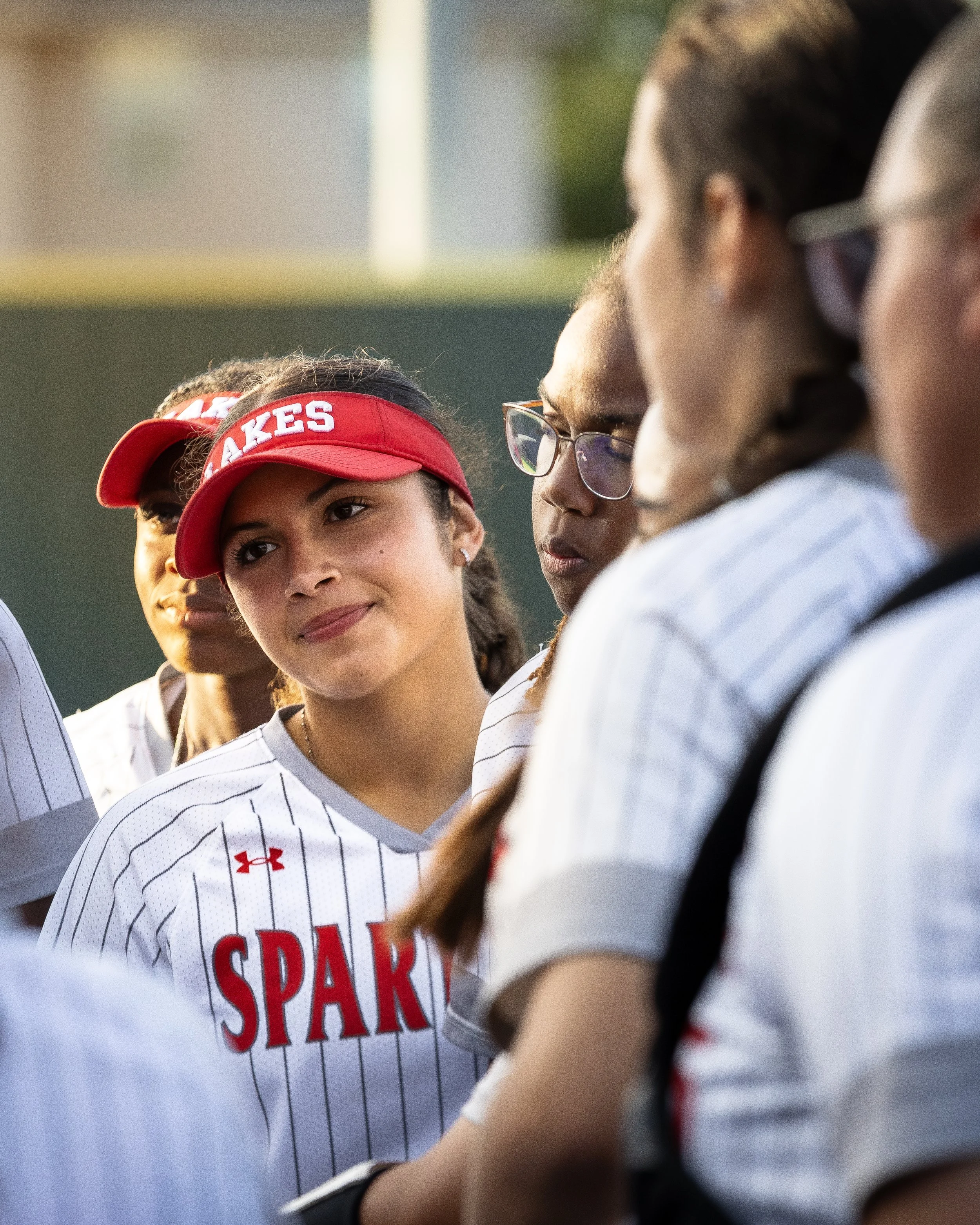 A group of young female softball players wearing white and red uniforms with 'SPARKS' written on the front, gathered together outdoors. One player is wearing a red visor.
