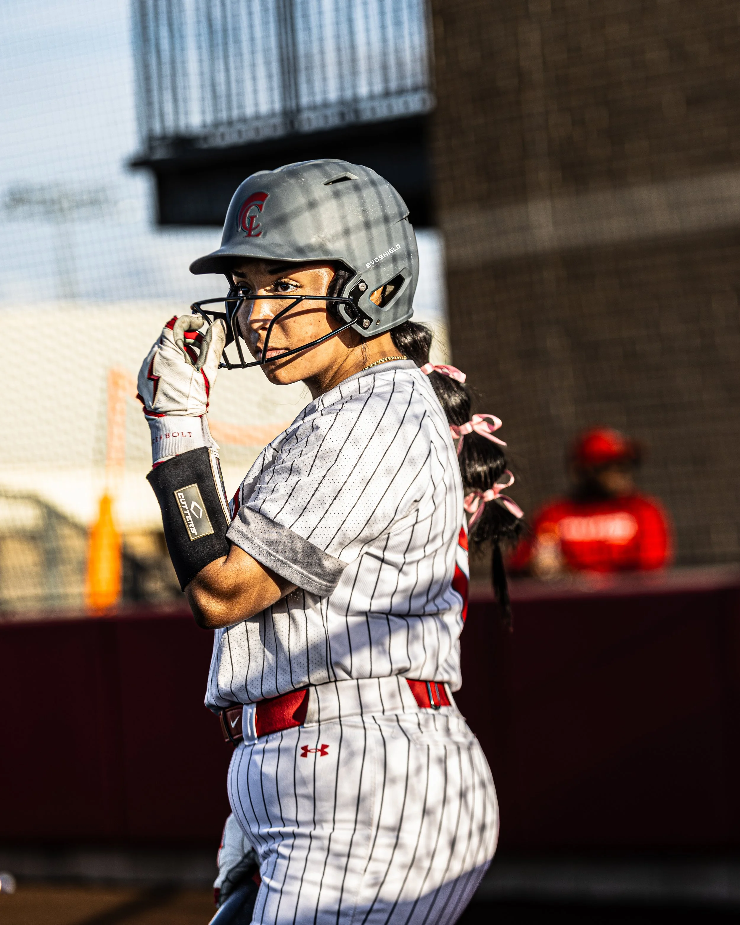 A female baseball player wearing a gray helmet with a red logo, pinstriped uniform, and batting gloves, standing on a baseball field, adjusting her helmet.