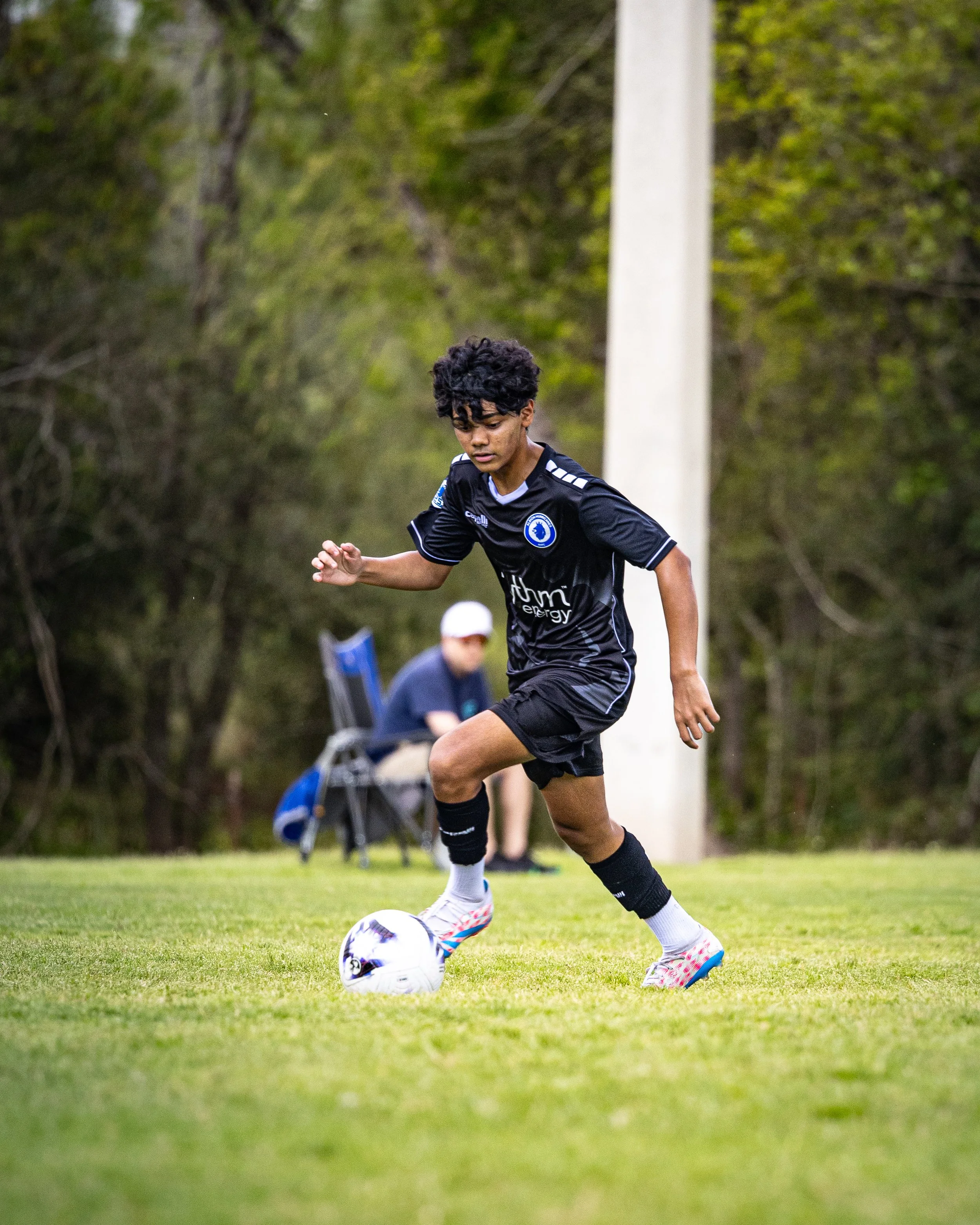 A young male soccer player in black uniform dribbling a white soccer ball on a grassy field during a game, with a person sitting in the background.