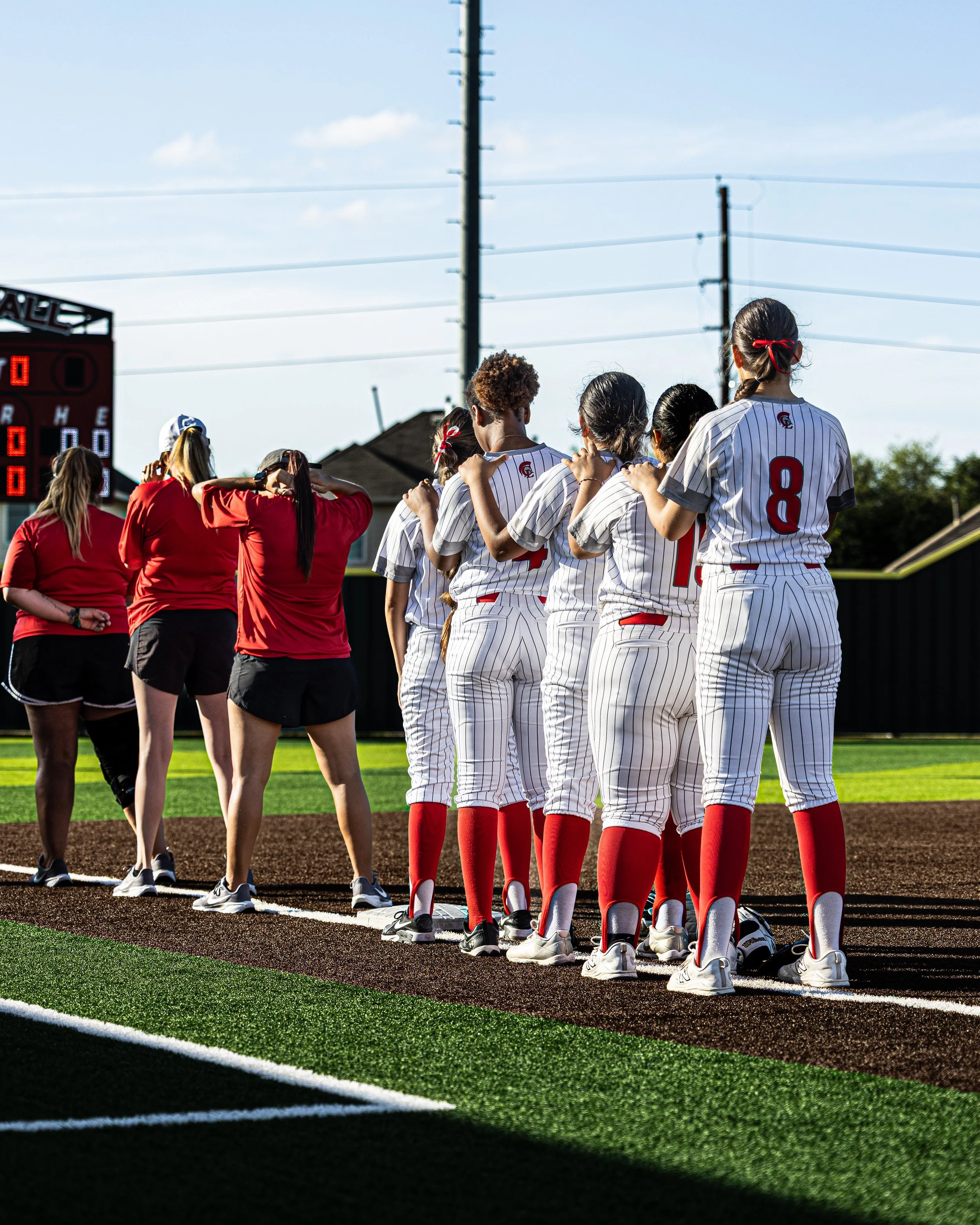 A group of female softball players in white pinstriped uniforms with red socks and cleats standing in a row on the field, with their hands on each other's shoulders, facing towards the left. Two other women in red shirts and black shorts are standing