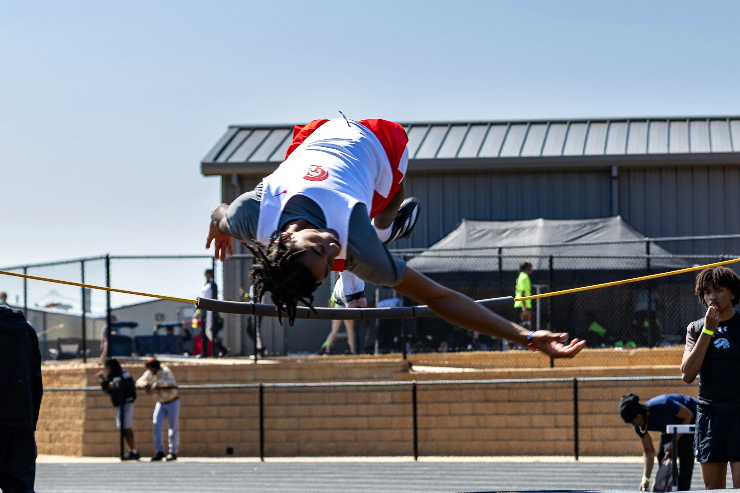 A male athlete with dreadlocks competing in a pole vault event, clearing the bar in an outdoor track and field stadium. Several people are watching in the background.