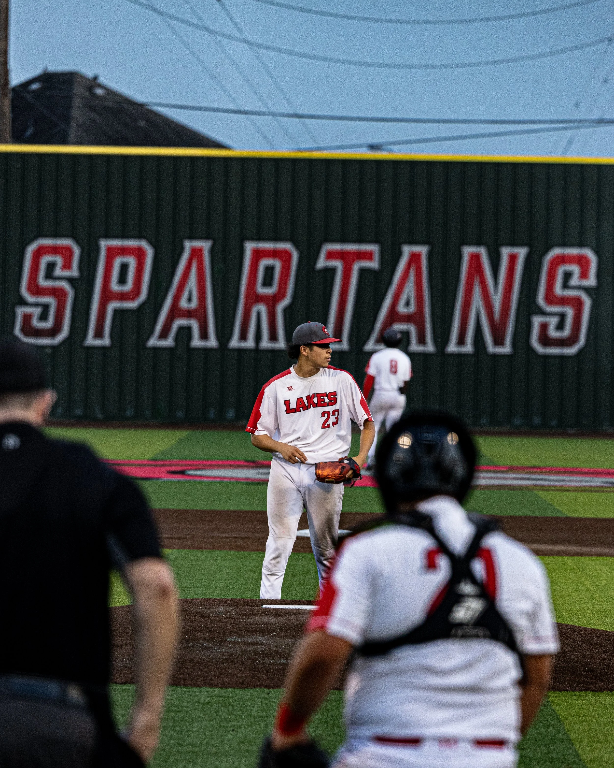 A baseball pitcher in a white and red uniform stands on the mound, preparing to pitch, with the word 'SPARTANS' on a wall behind him. Two other players and a coach or umpire are nearby on the field.