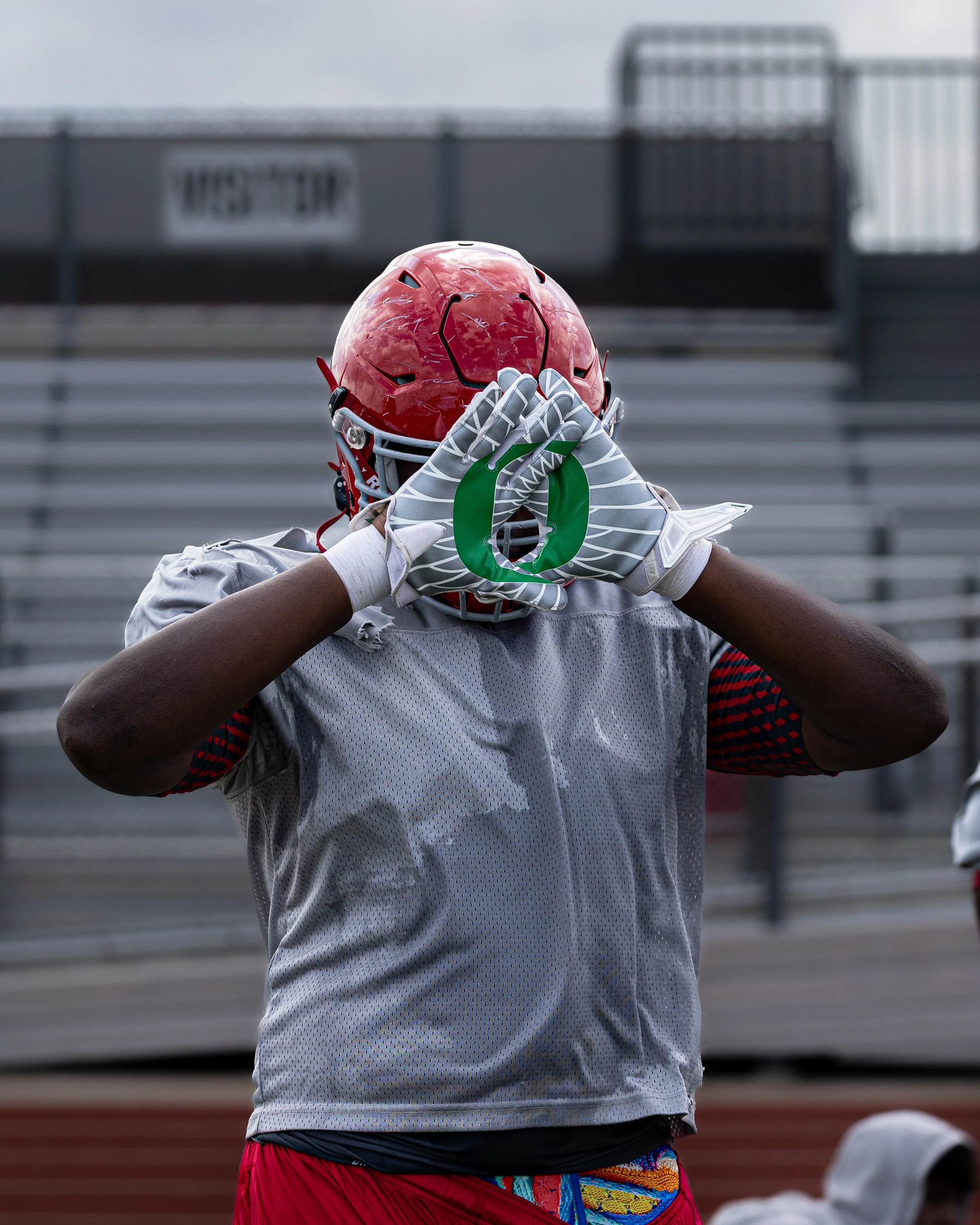 American football player wearing a red helmet, white jersey, and colorful shorts, holding hands in a diamond shape near his face on a football field.