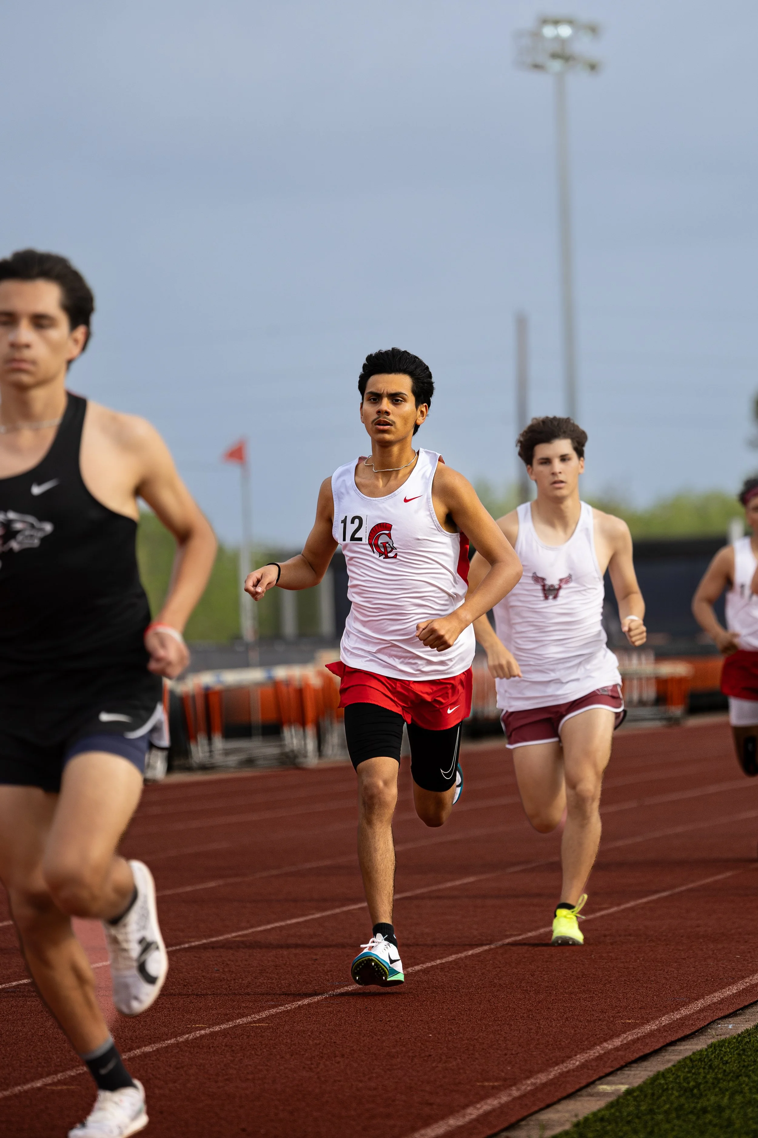 Group of young athletes running on a track during a race, with focus on a male runner wearing a white and red uniform with the number 12.