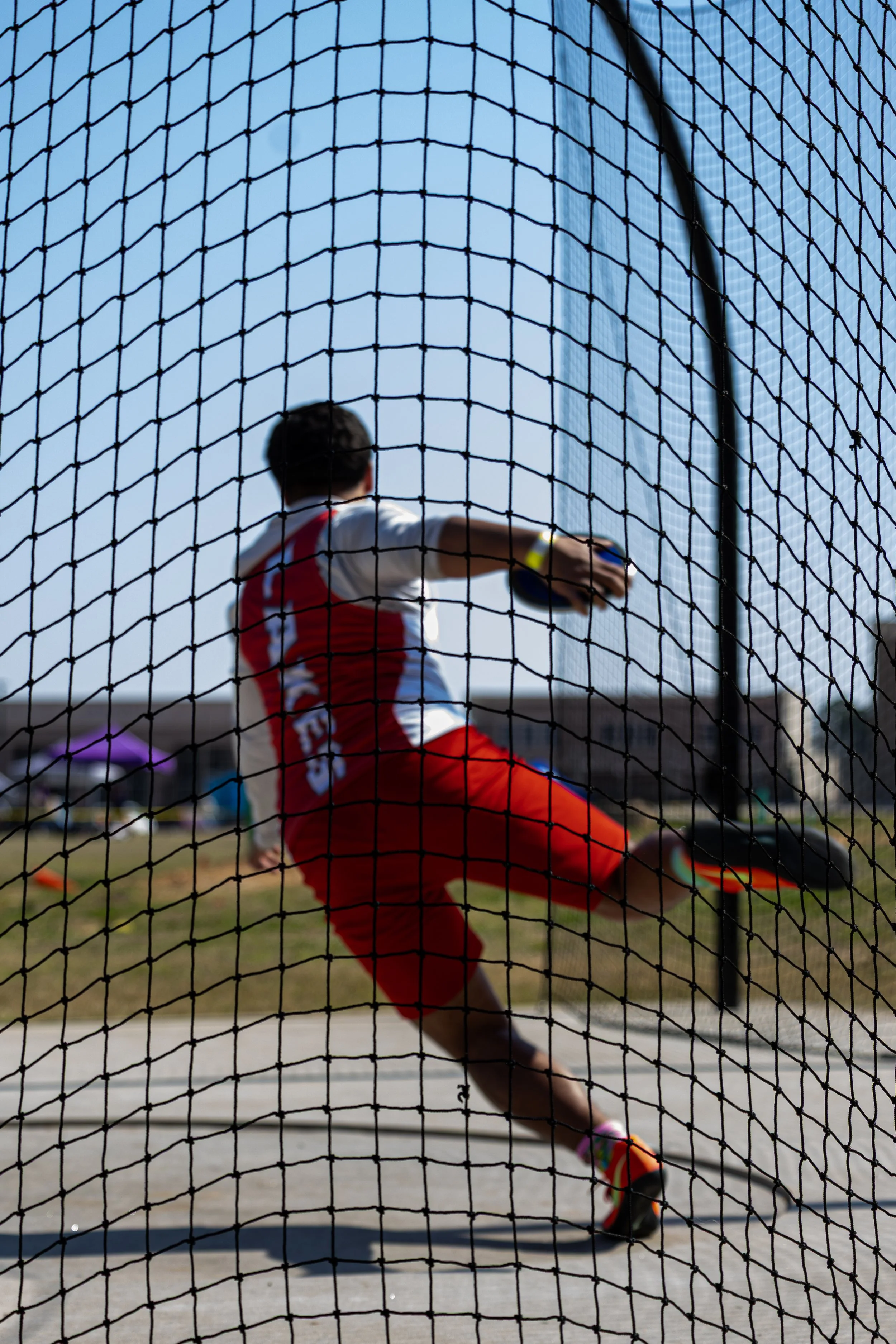 A male athlete in a red and white sports uniform throwing a shot put during a track and field event.