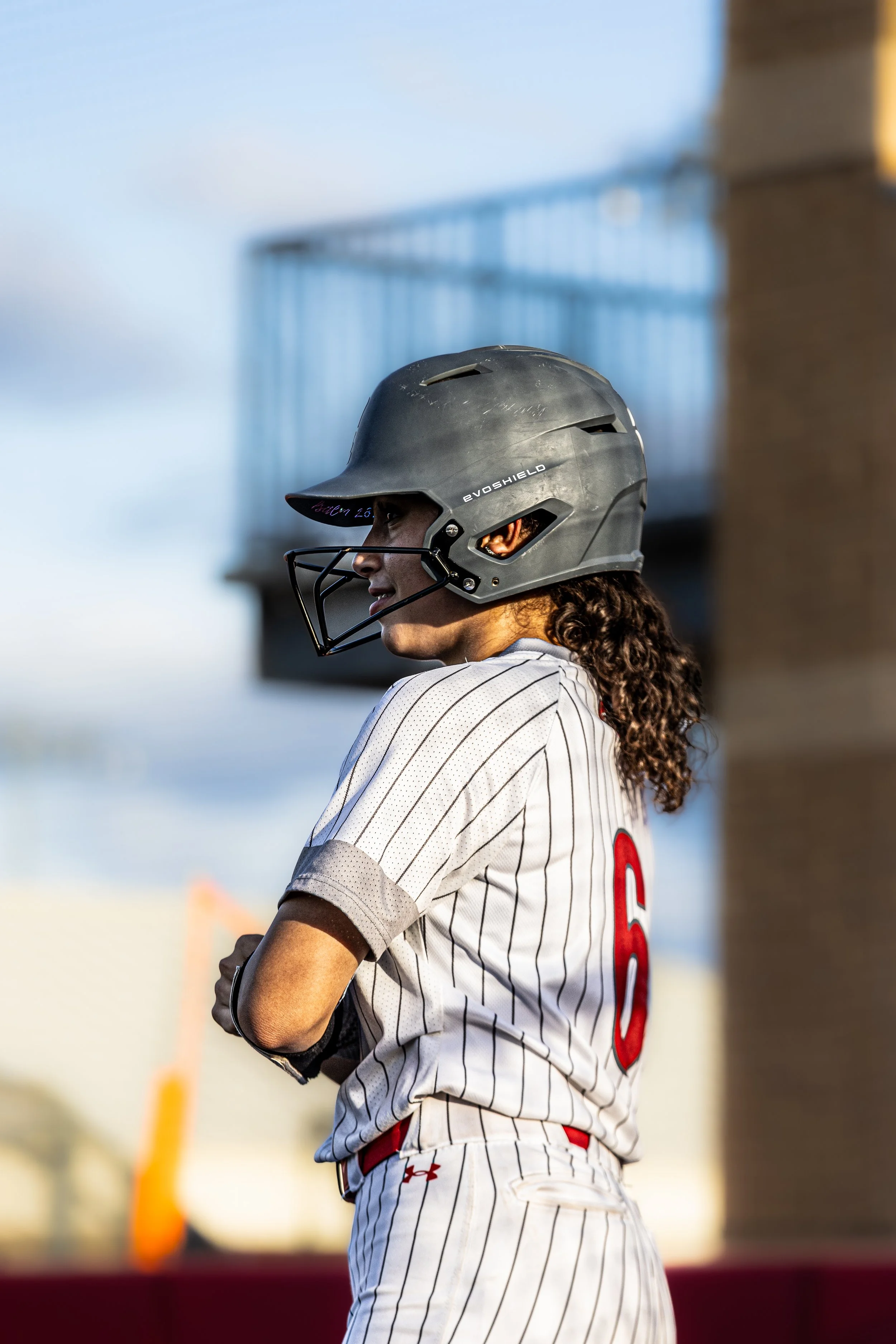 A female baseball player wearing a black helmet with a faceguard and a white pinstriped uniform with the number 6 on the back, standing outdoors.