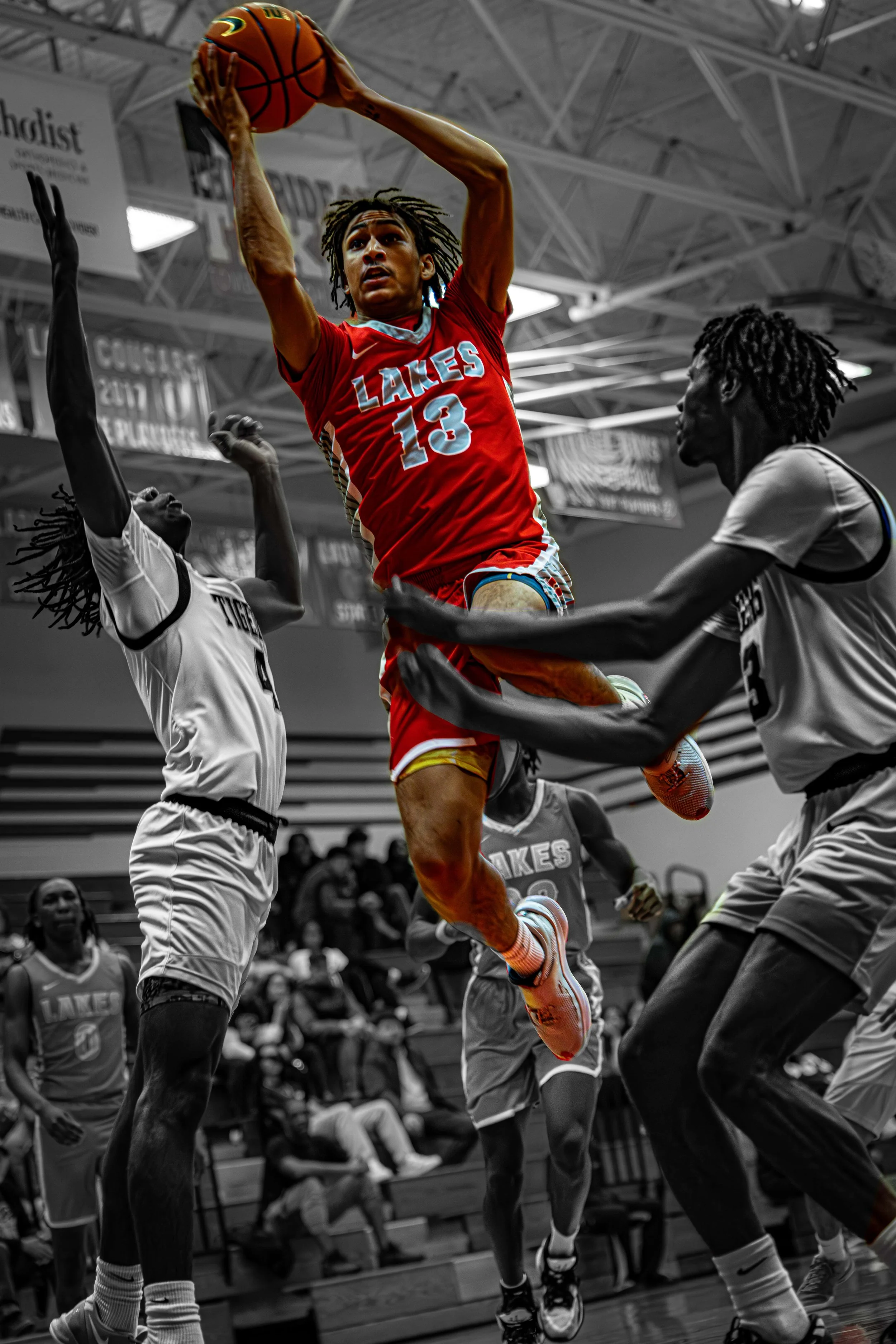 A basketball player wearing a red uniform with the number 13, jumping to make a shot while being guarded by two opponents in white uniforms in a gymnasium.