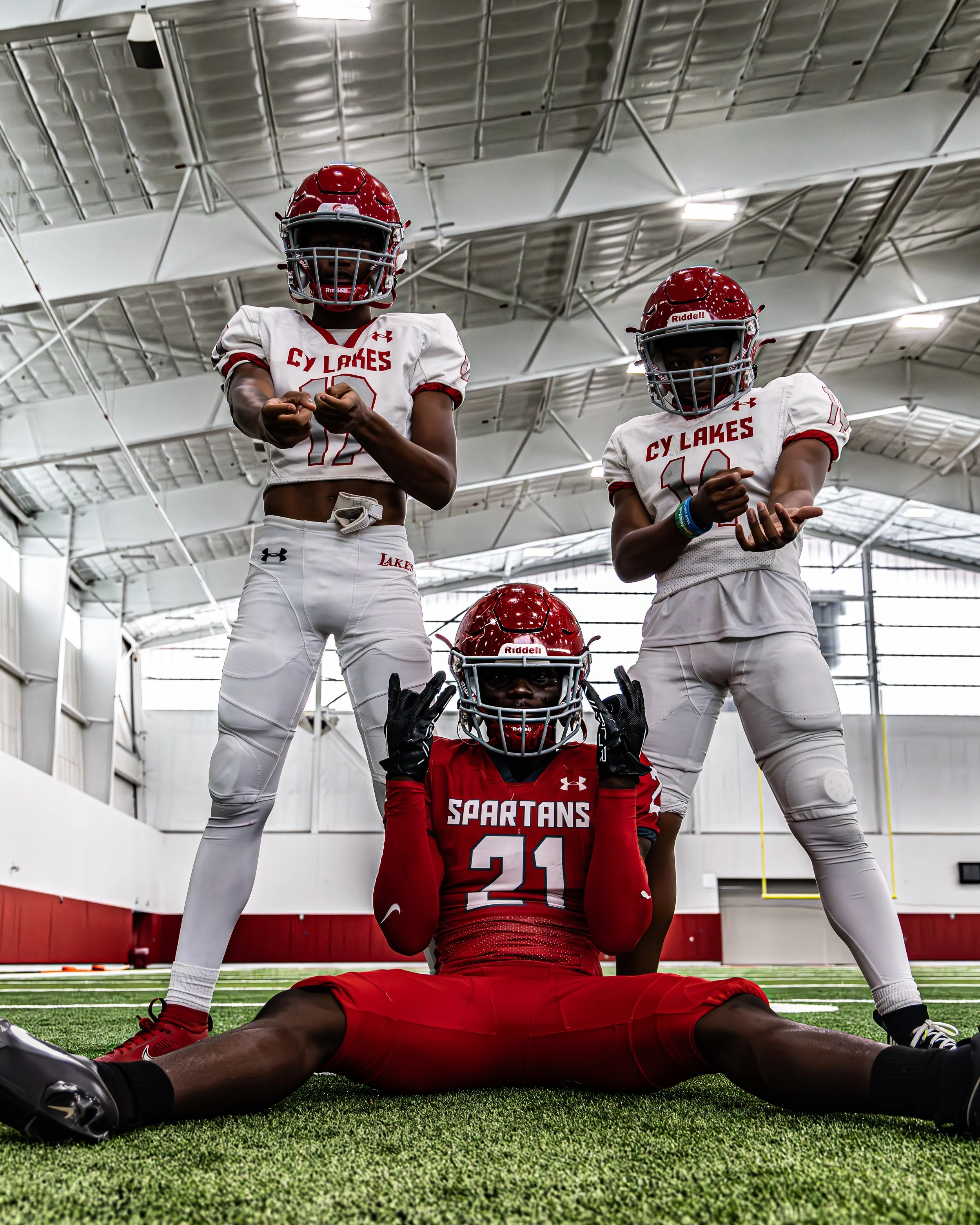Three football players in uniforms and helmets inside an indoor football field, one sitting on the ground making a peace sign and two standing behind with their fists clenched.
