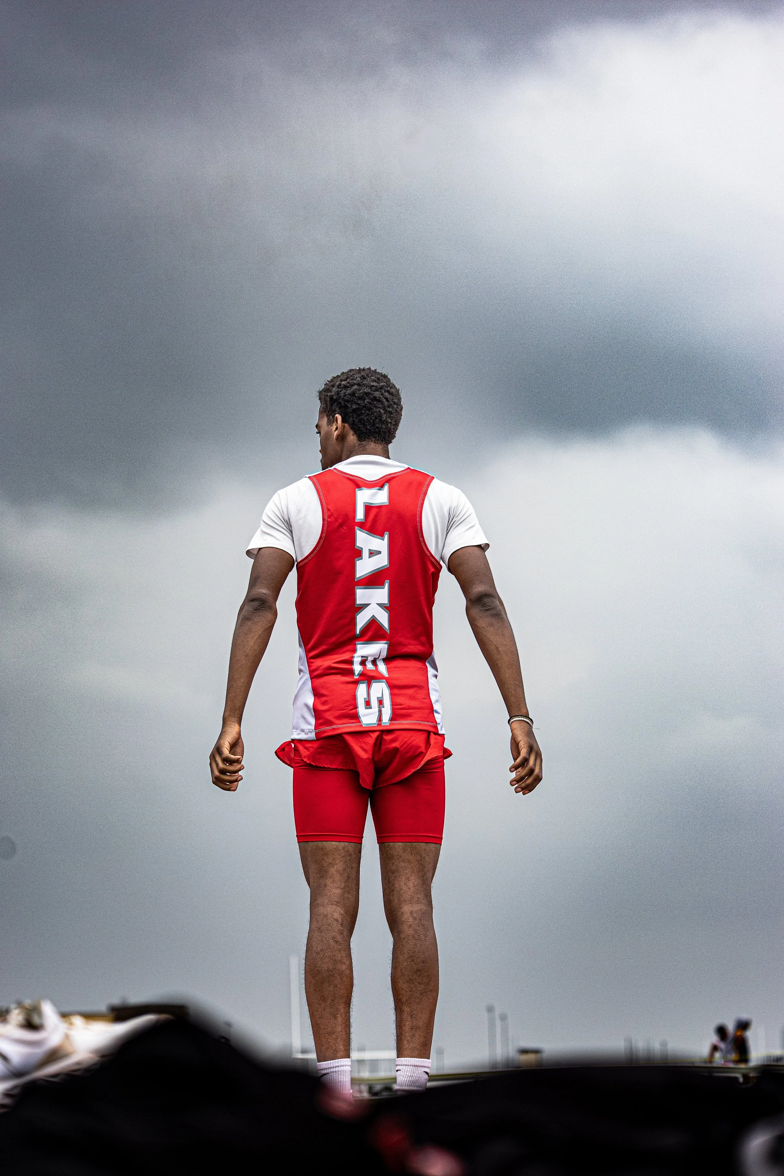 Back view of a male athlete in a red and white team uniform with 'LAKES' written on the back, standing outdoors under dark, cloudy sky.