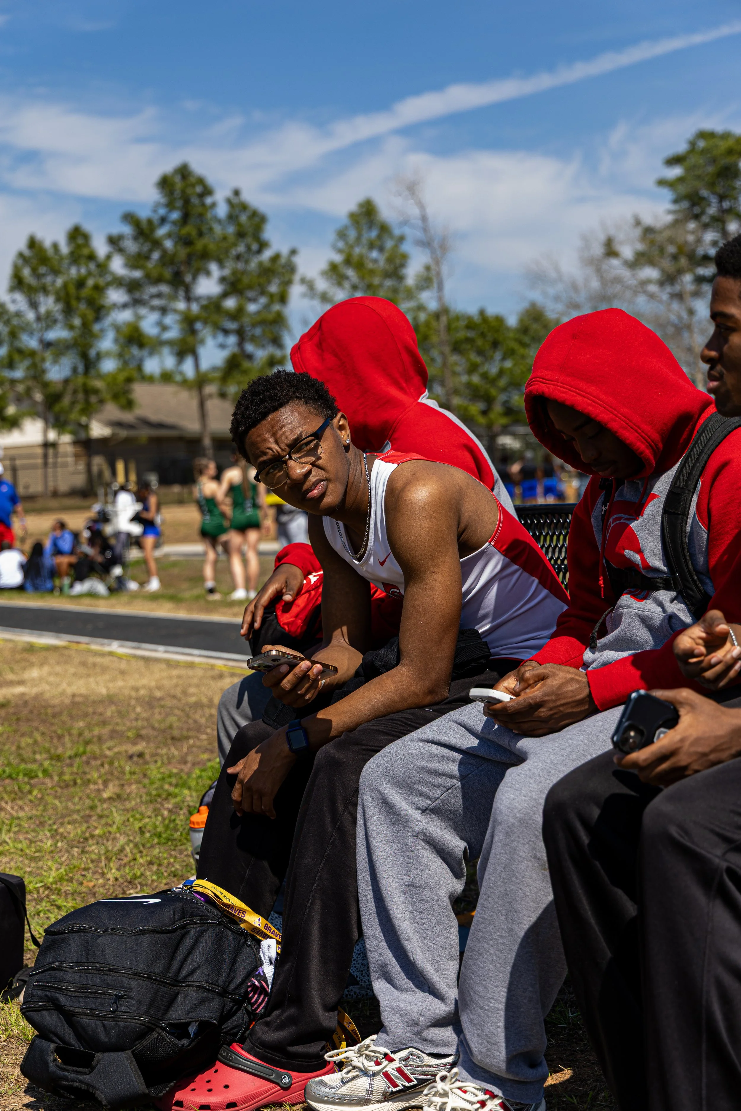 A young male athlete with glasses and a sleeveless shirt sitting on a bench at a track meet, looking at the camera. He is surrounded by other young men in red hoodies, some with their heads down. In the background, spectators and other athletes are v
