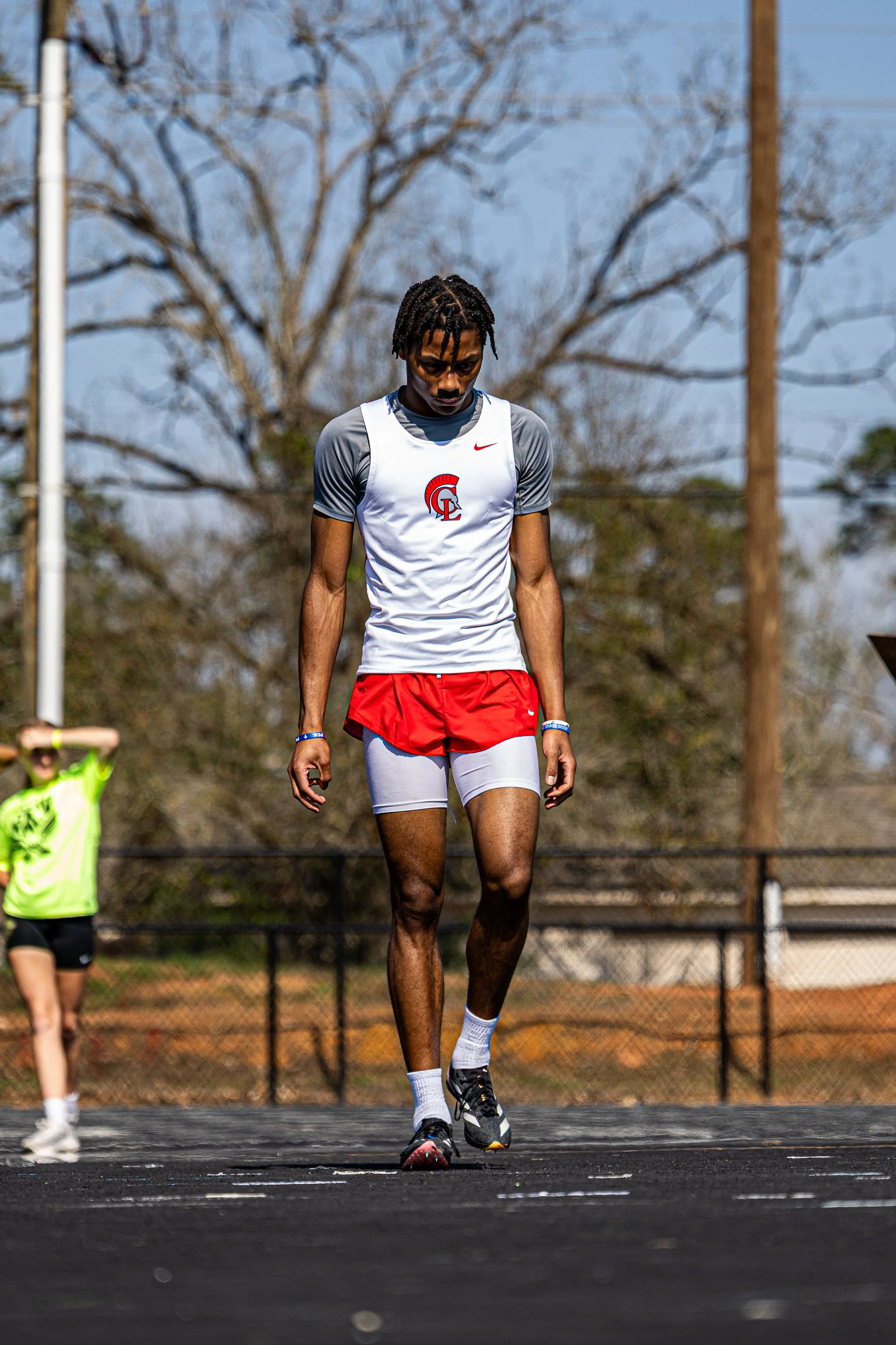 A male athlete with dreadlocks standing on a track wearing a gray and white Nike tank top with a logo, red shorts, and black running shoes, with a woman in a yellow shirt in the background and leafless trees and power lines behind.