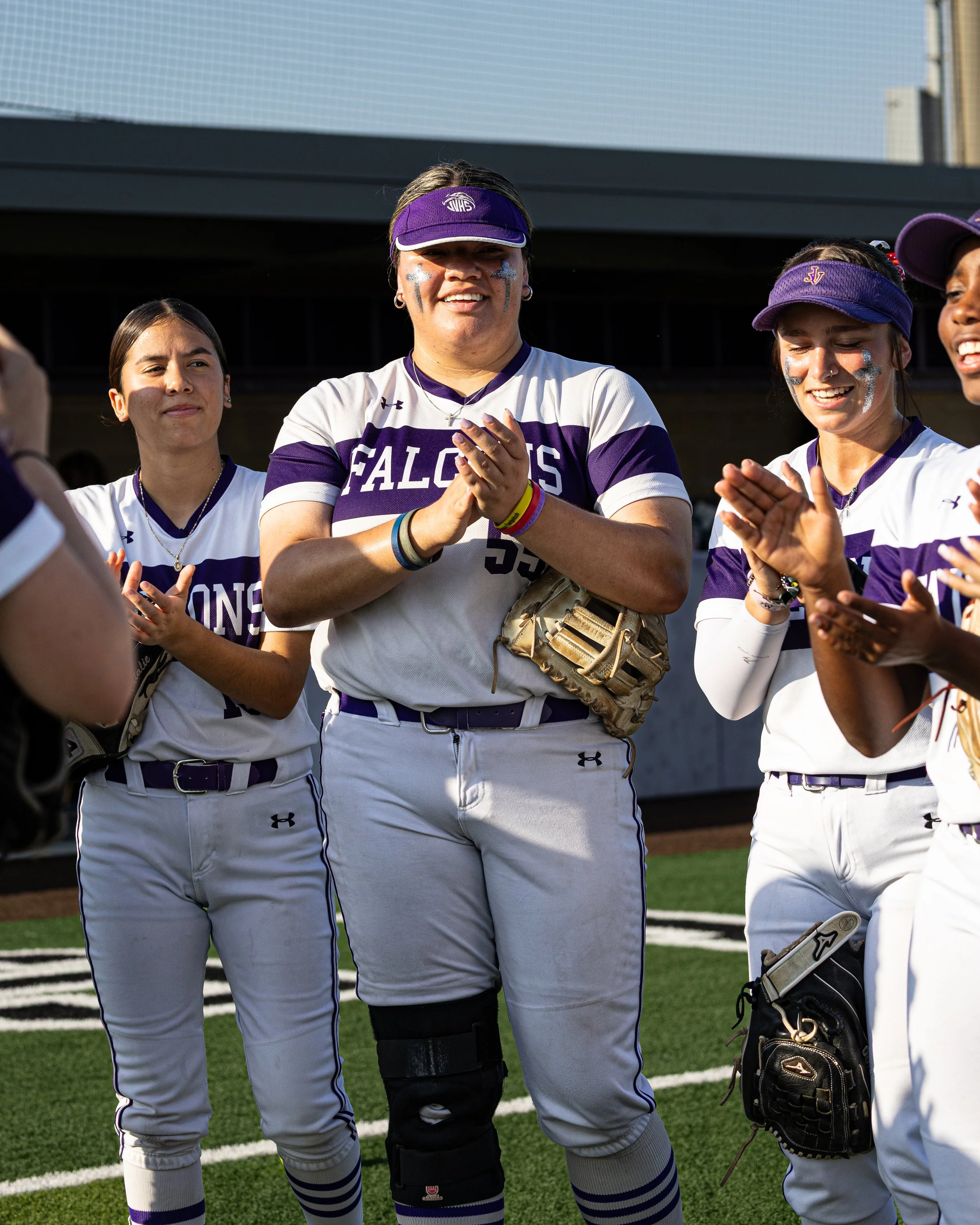 A group of female softball players in uniform on the field, smiling and clapping during a game or practice.
