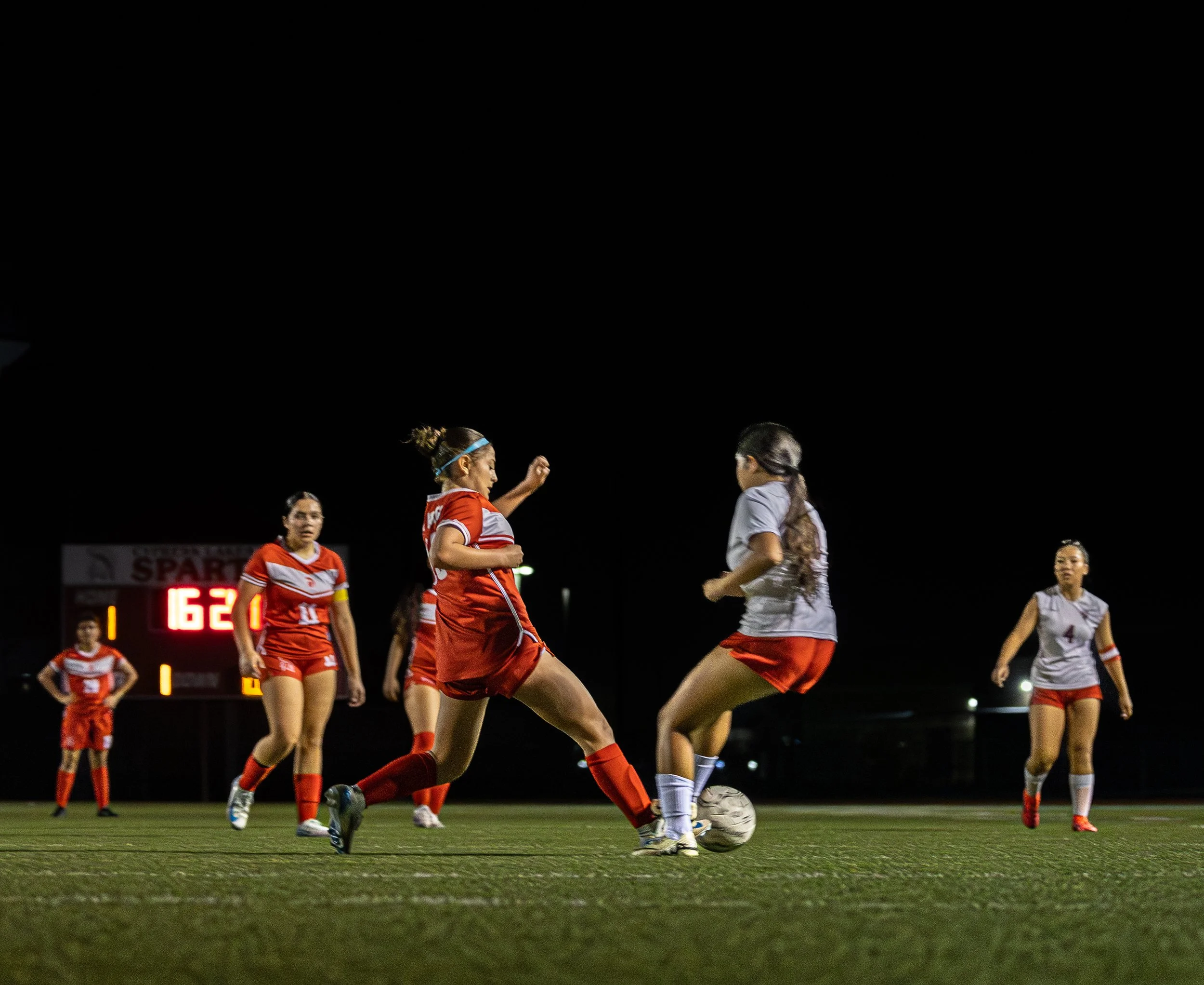 Soccer players at night on a field, two players in red and white uniforms competing for the ball, with others watching in the background.
