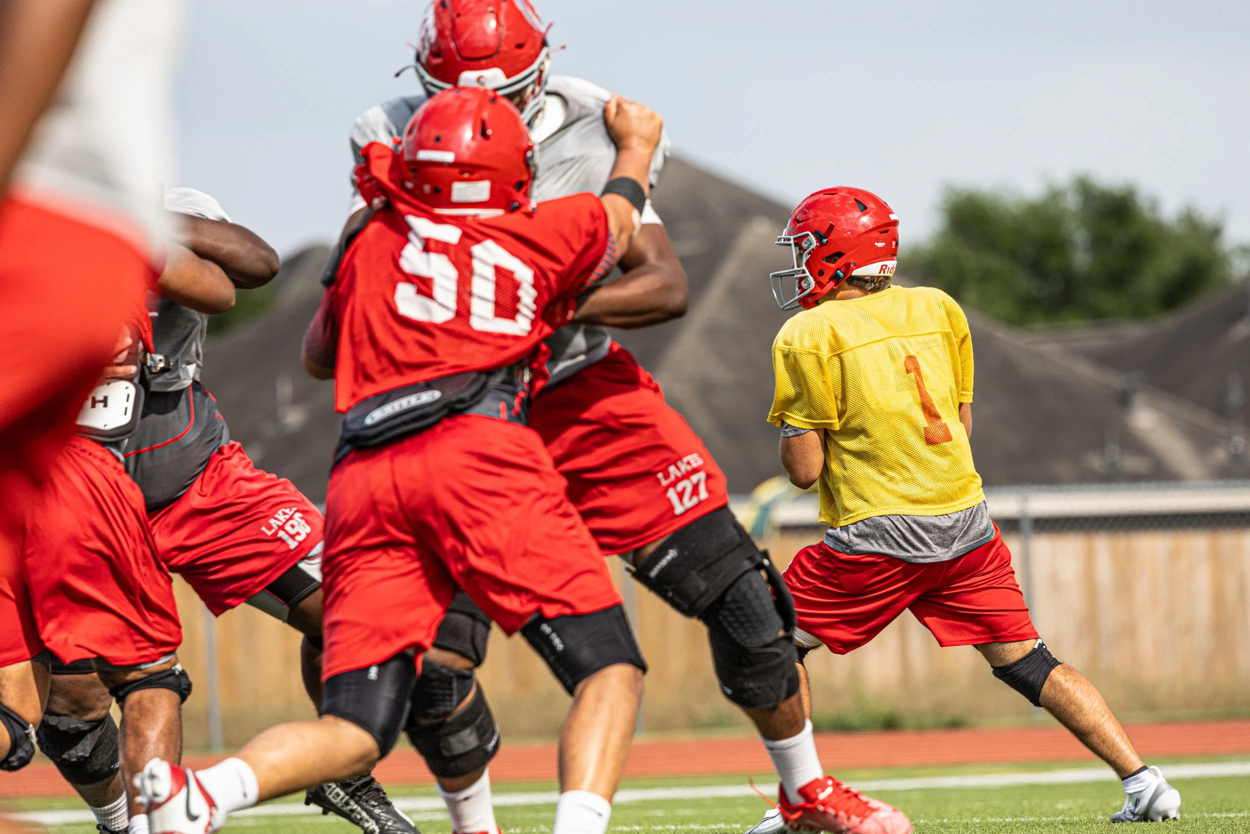 Young football players practice on the field, with a quarterback in a yellow jersey and red helmet ready to throw. Other players in red uniforms are blocking or preparing for the play.