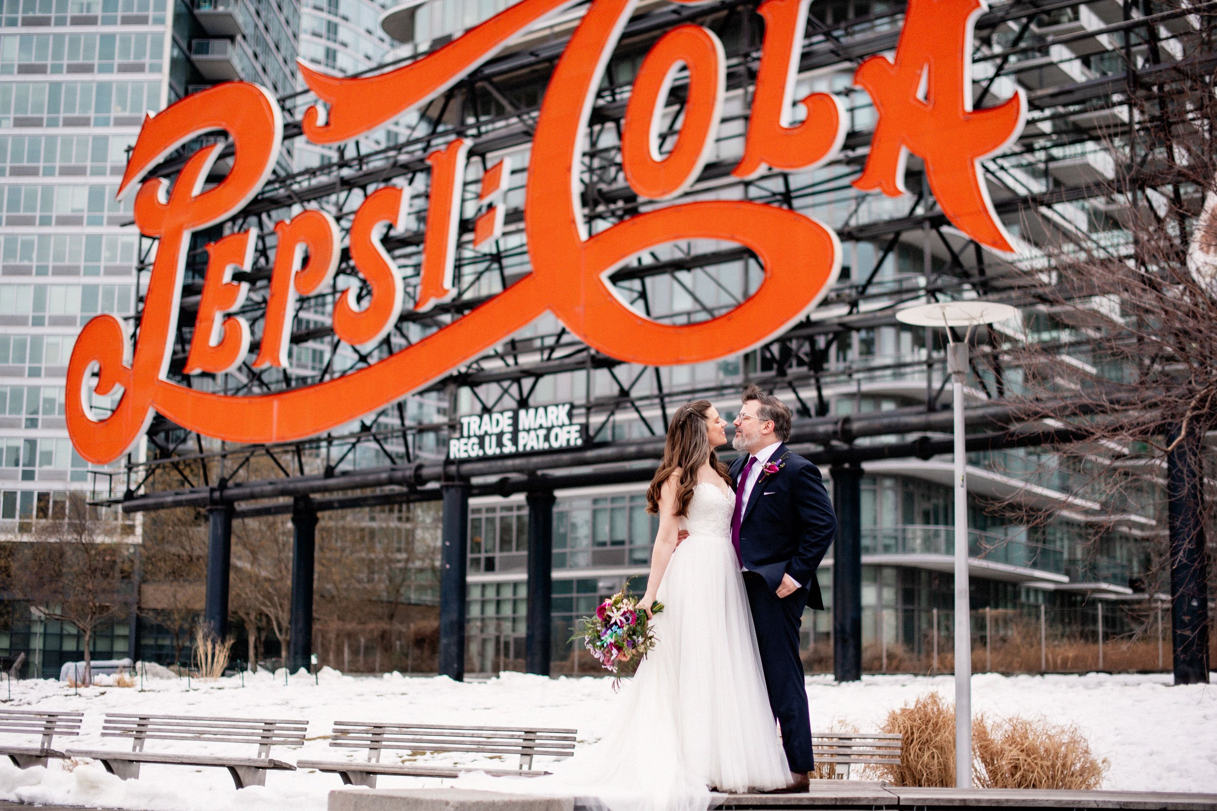 A newlywed couple standing in front of a large Pepsi-Cola sign as they are about to kiss