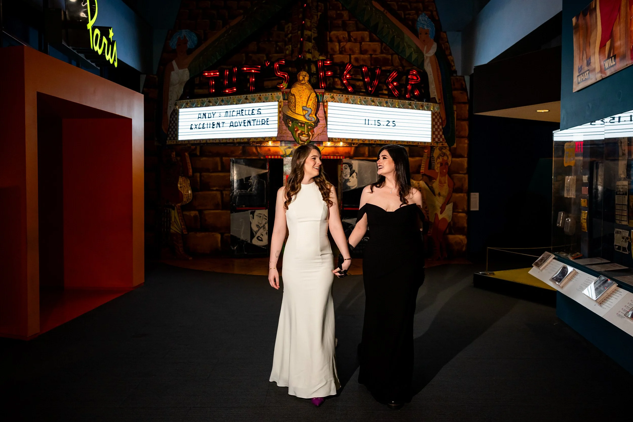 A newlywed couple holding hands and smiling at each other as they walk out of a theater