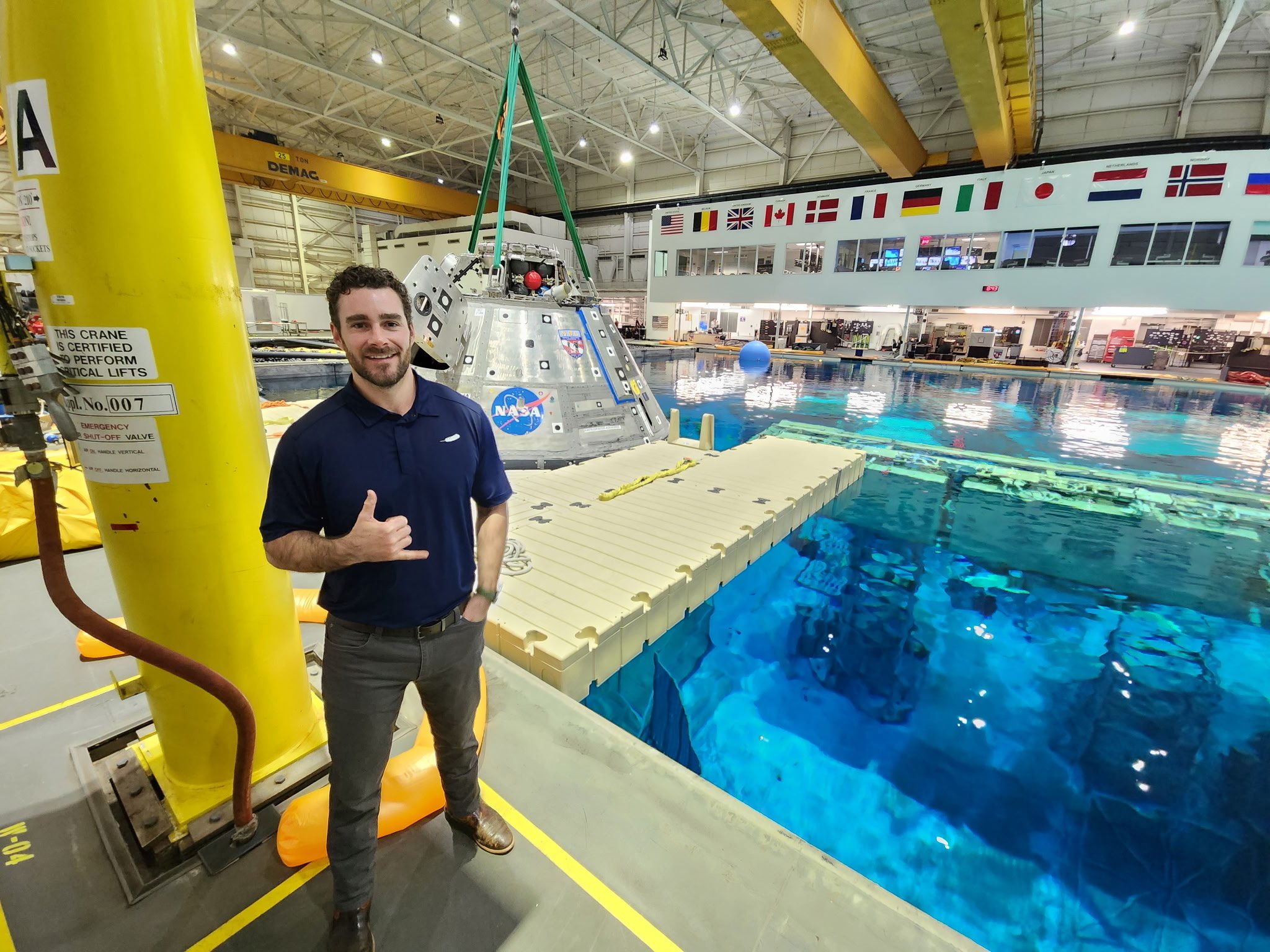 A man smiling and giving a shaka sign in front of a NASA lunar module simulator inside a large training facility with a swimming pool, flags from different countries, and various equipment in the background.