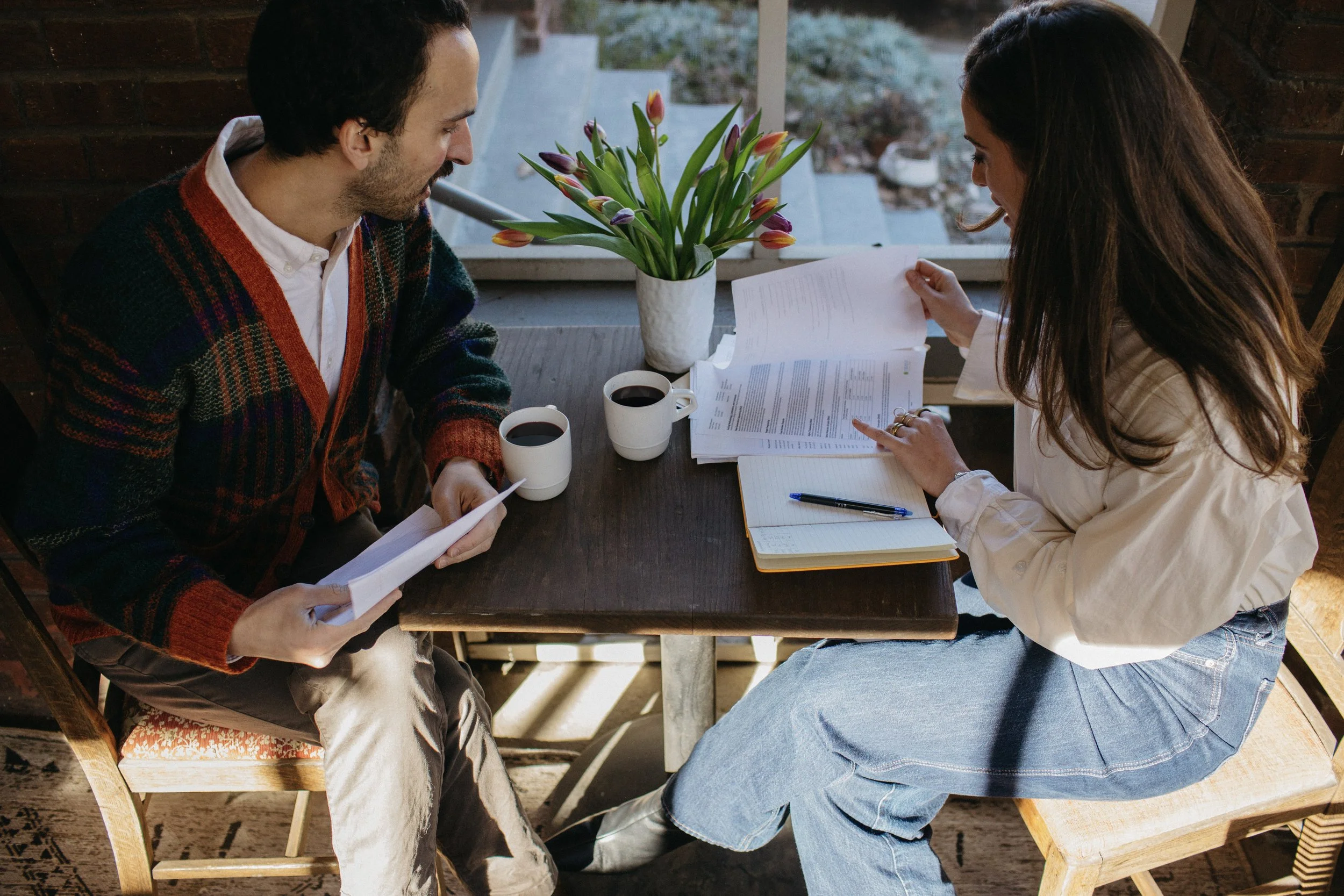 A man and woman sitting at a wooden table, reviewing documents with two cups of coffee and a vase of tulips between them. They are in a cozy space with natural light coming from a window behind.
