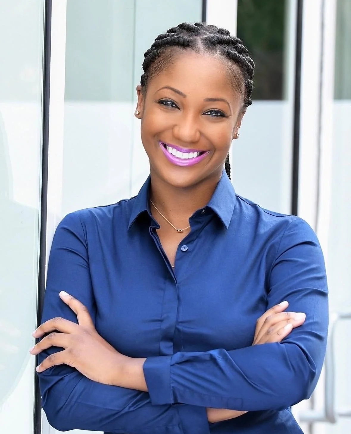 Smiling woman with braided hair wearing a blue shirt, standing outdoors with glass walls in the background.