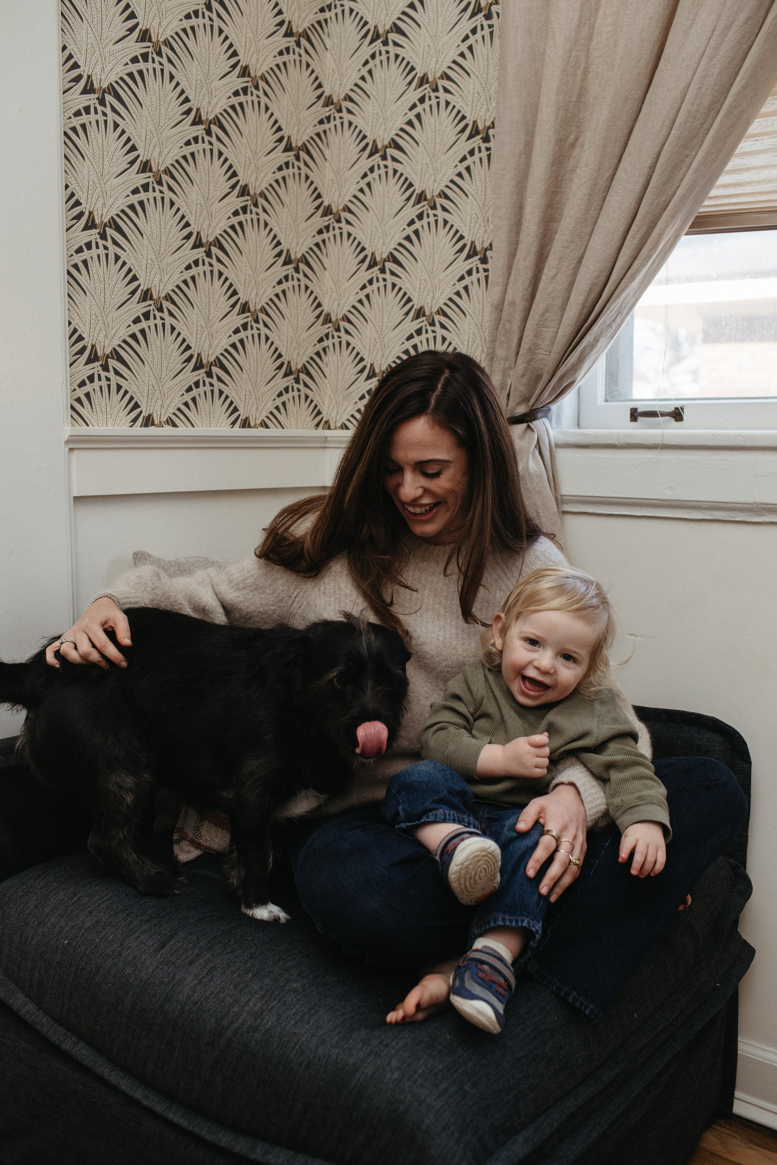 A woman and a young boy sitting on a black couch in a room with patterned wallpaper and beige curtains. The woman is smiling, and the boy is sitting on her lap, also smiling. A black puppy with its tongue out is on the couch, close to the woman.