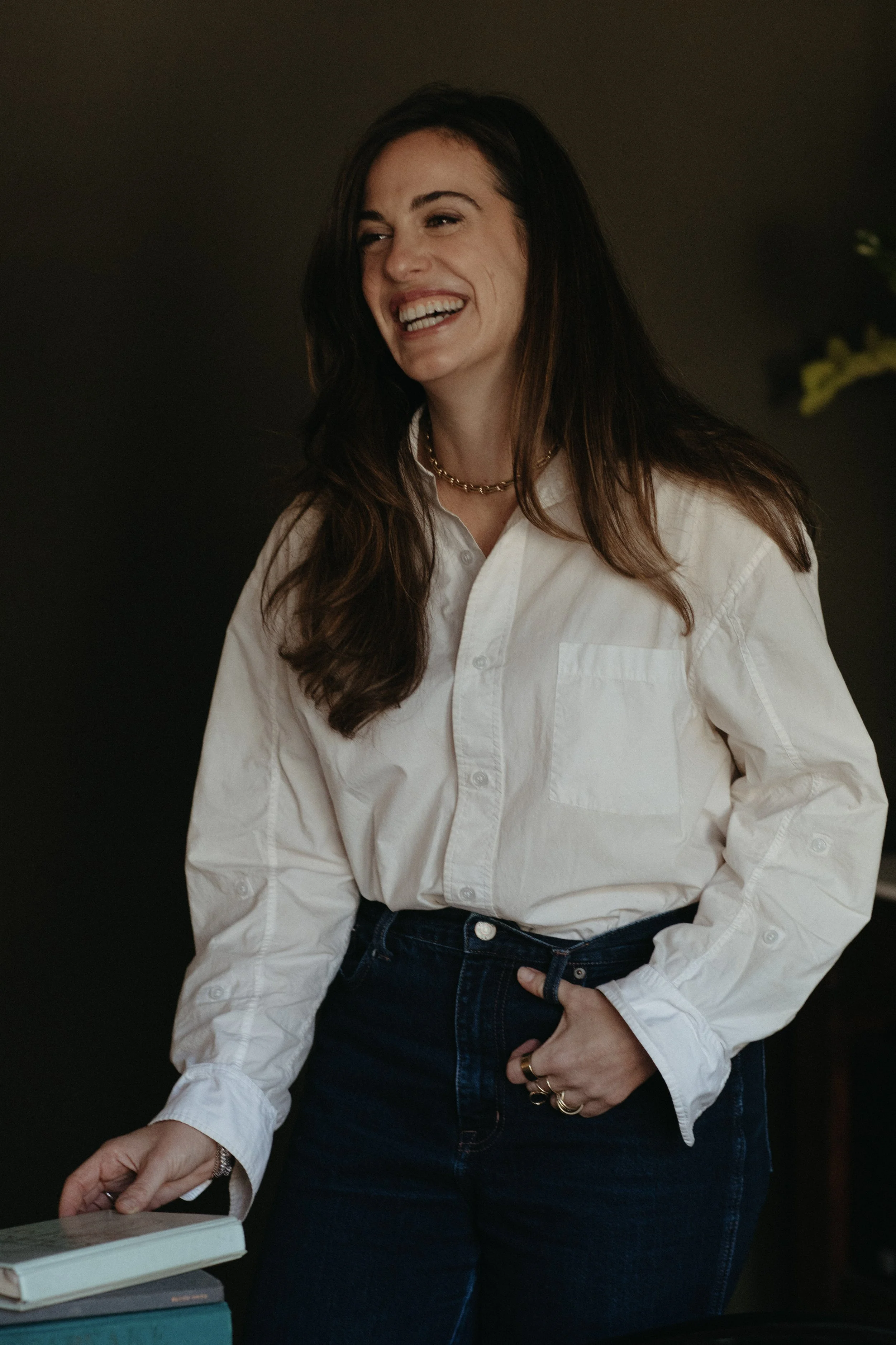 A woman with long brown hair smiling and wearing a white button-up shirt and dark jeans.