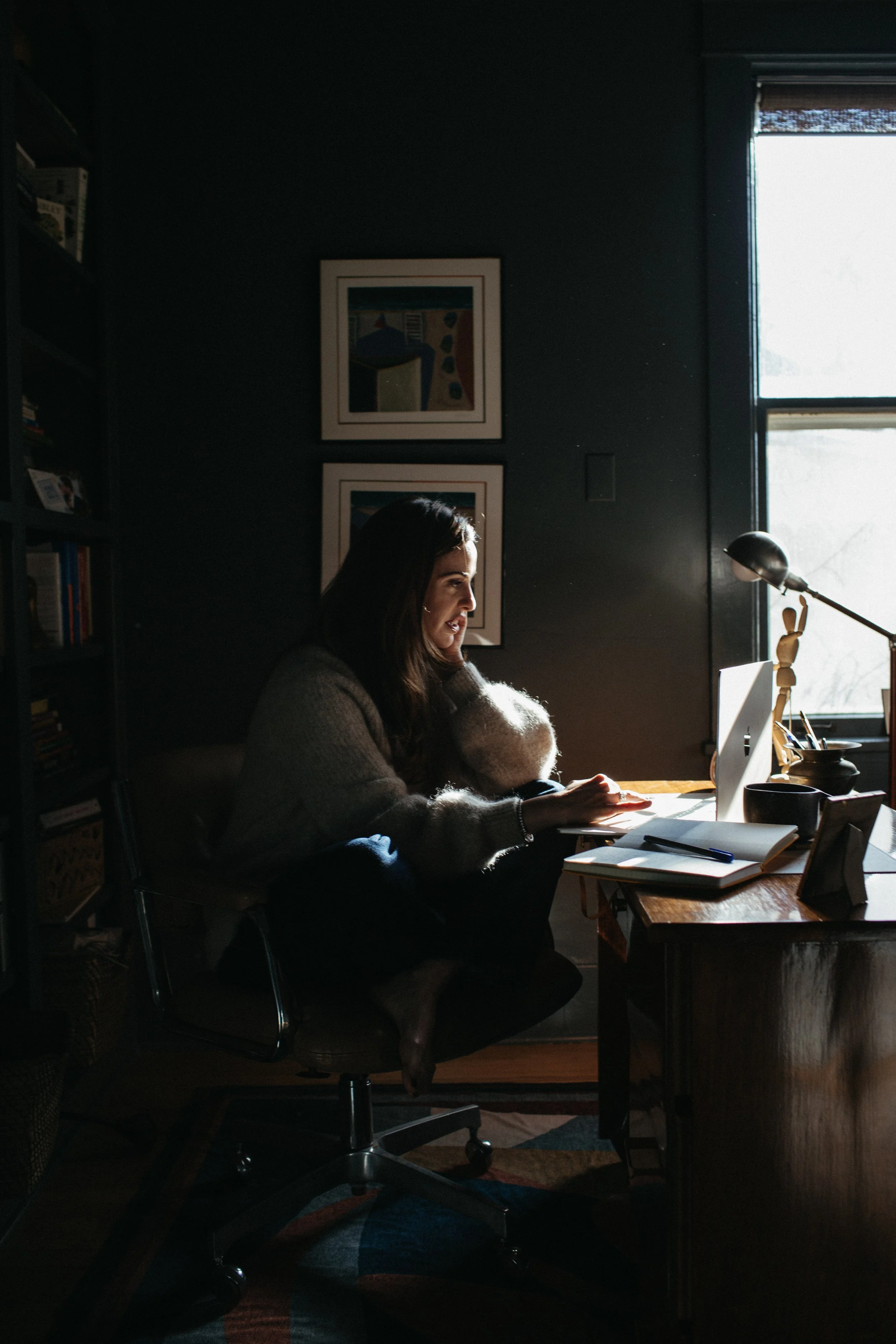 A woman working or studying at a desk in a dark room with sunlight coming in through a window on the right side.