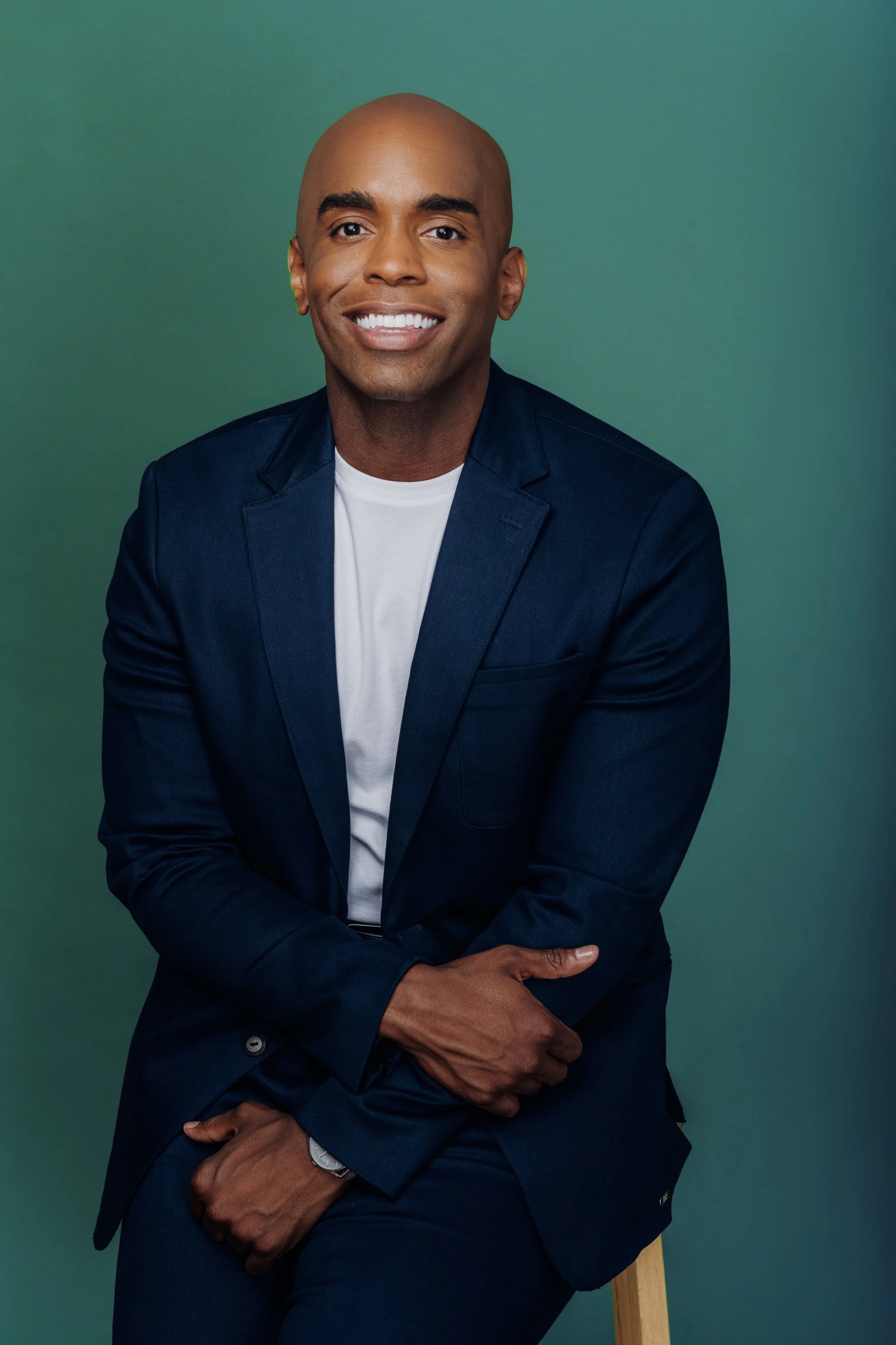 A man in a navy blue blazer and white shirt sitting on a stool against a green background, smiling at the camera.