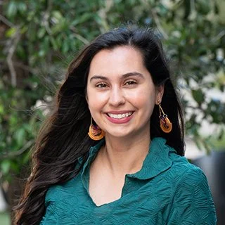 Smiling woman with long dark hair, wearing earrings and a teal top, standing outdoors with greenery in the background.