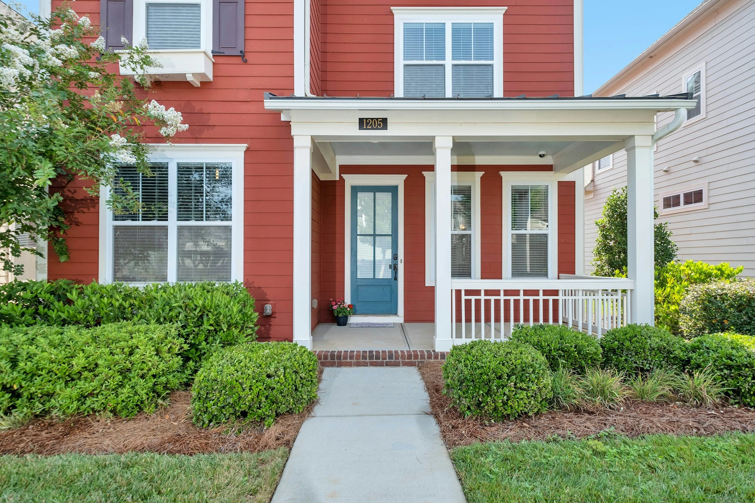 Front porch of a red house with white trim, blue door, and a small garden with green bushes and a flower pot.