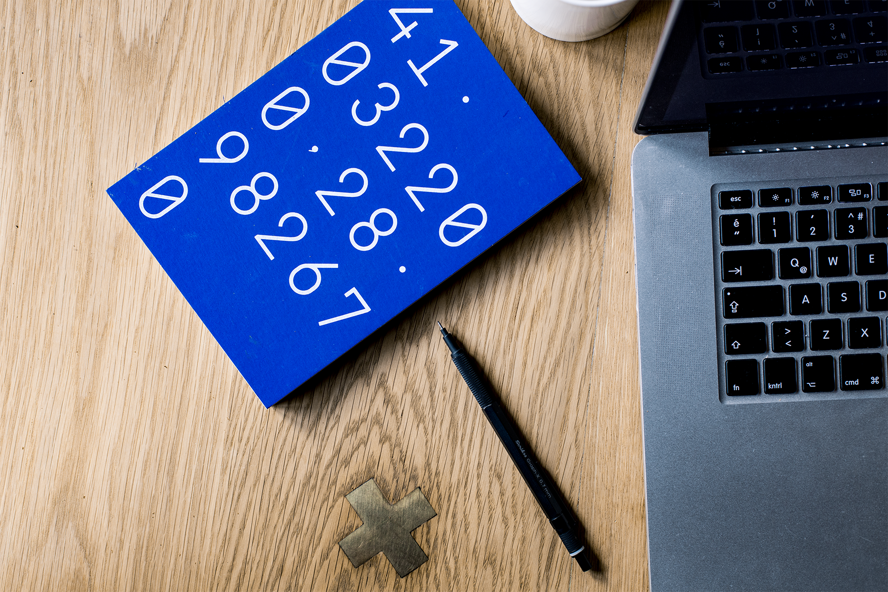 A wooden desk with a blue brain teaser puzzle, a black pen, a small metal cross, a coffee mug, and part of a silver laptop with a black keyboard.