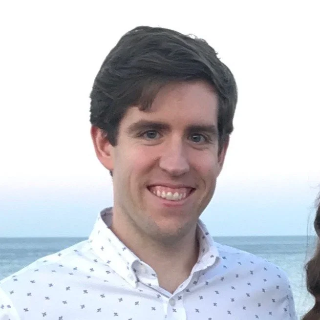 Young man with dark hair smiling outdoors at the beach with ocean in the background.