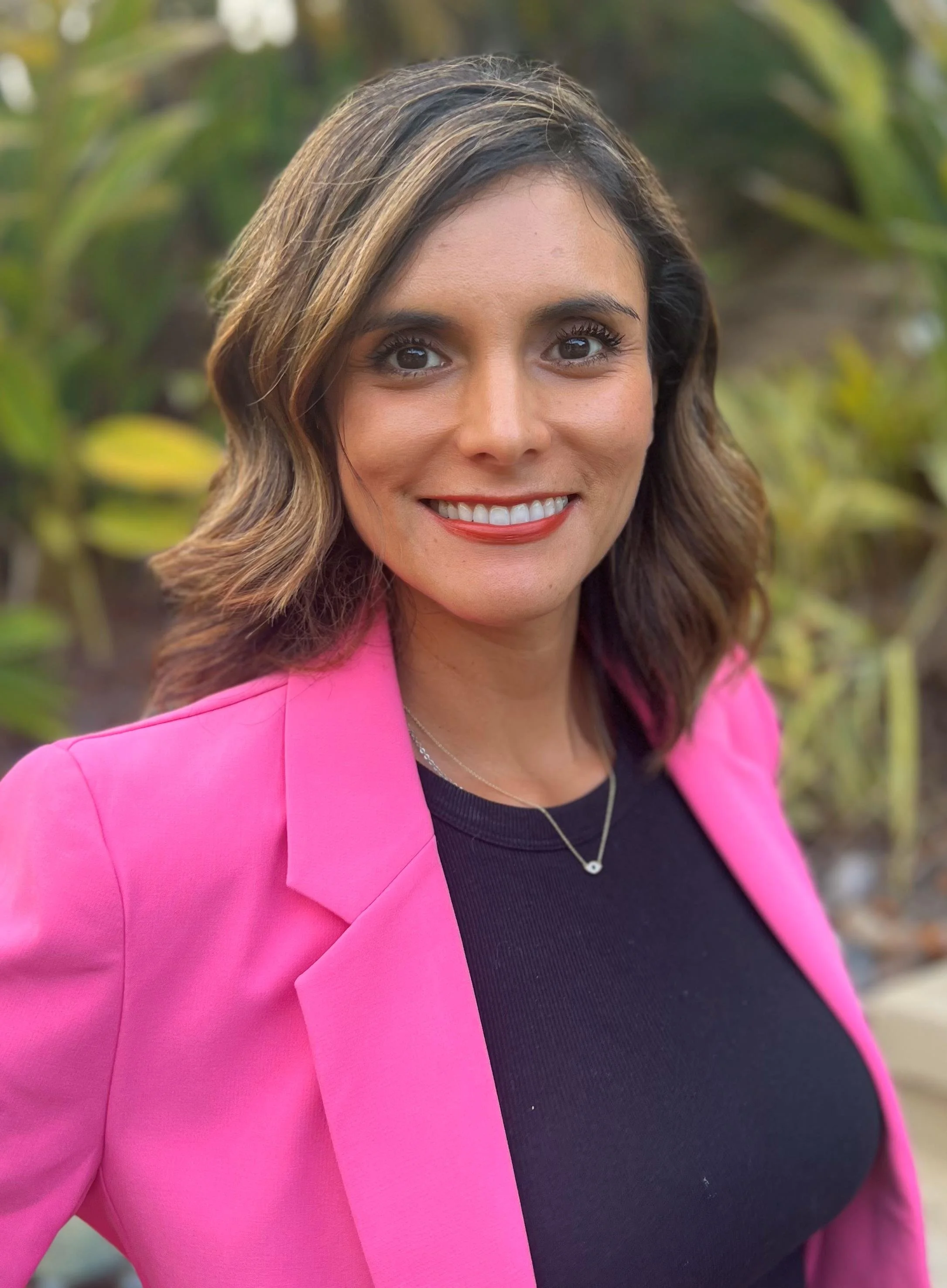 Close-up of a smiling woman with shoulder-length brown wavy hair, wearing a bright pink blazer and a black top, outdoors with green foliage in the background.