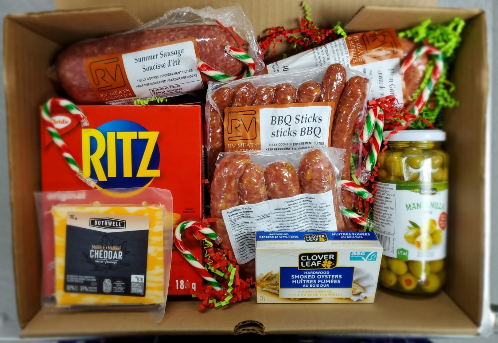 Gift gift box filled with assorted meats, cheese, snacks, and canned olives, including summer sausage, BBQ sticks, smoked oysters, cheddar cheese, and a jar of green olives, decorated with red and green paper shreds.