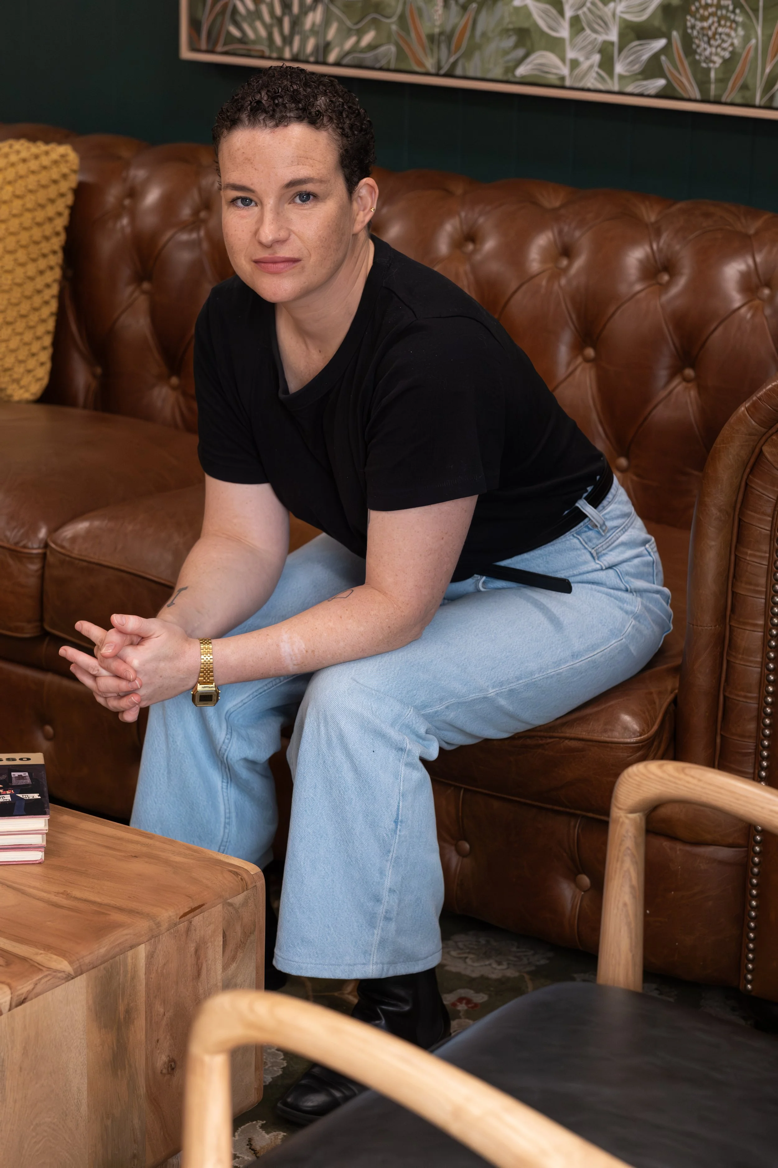 A person with short, curly hair and light skin, wearing a black T-shirt and light blue jeans, sitting on a brown leather sofa with their hands clasped, looking at the camera.