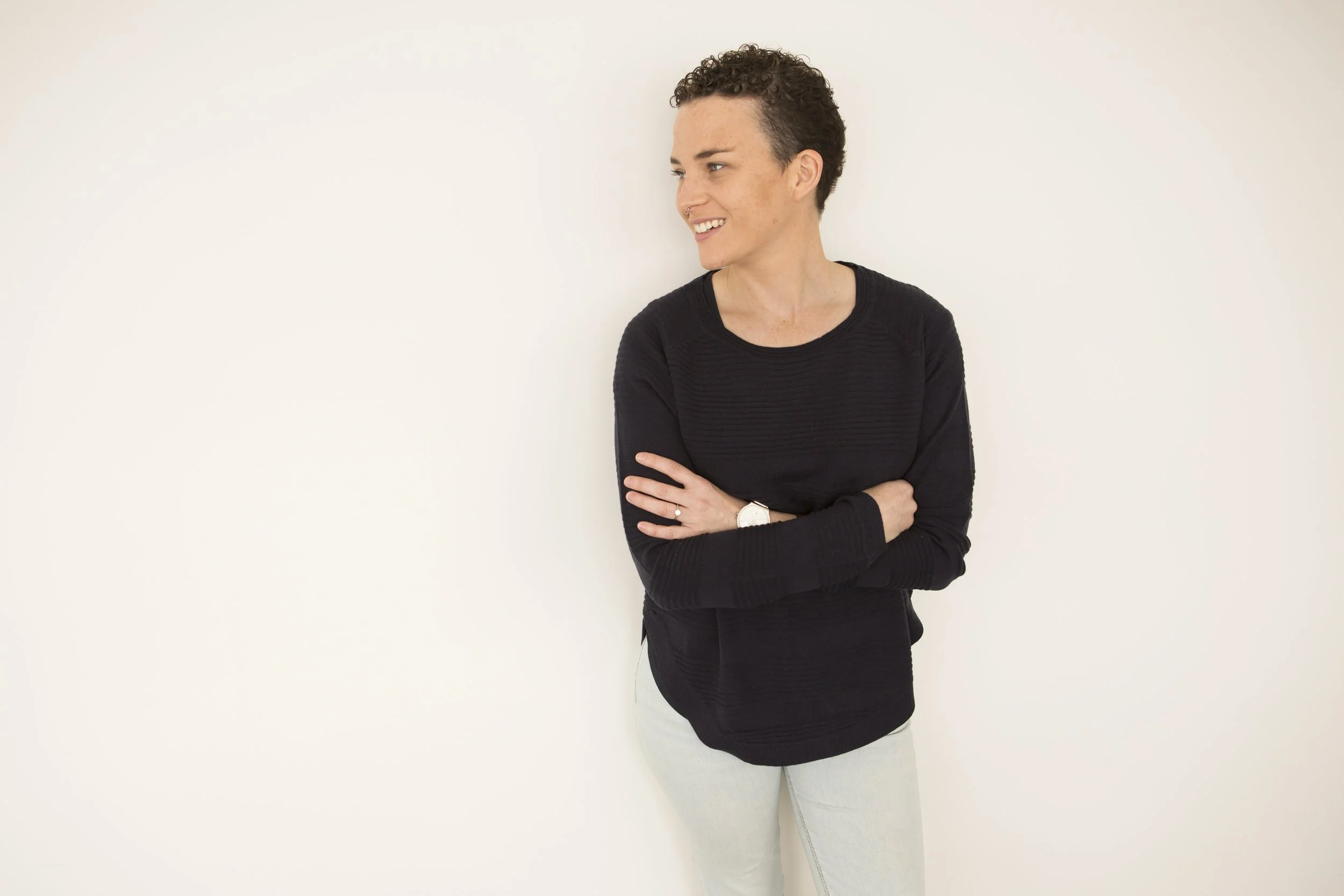 A woman with short curly hair, wearing a black long-sleeve shirt and light-colored pants, standing against a plain white wall with her arms crossed, looking to her left and smiling.