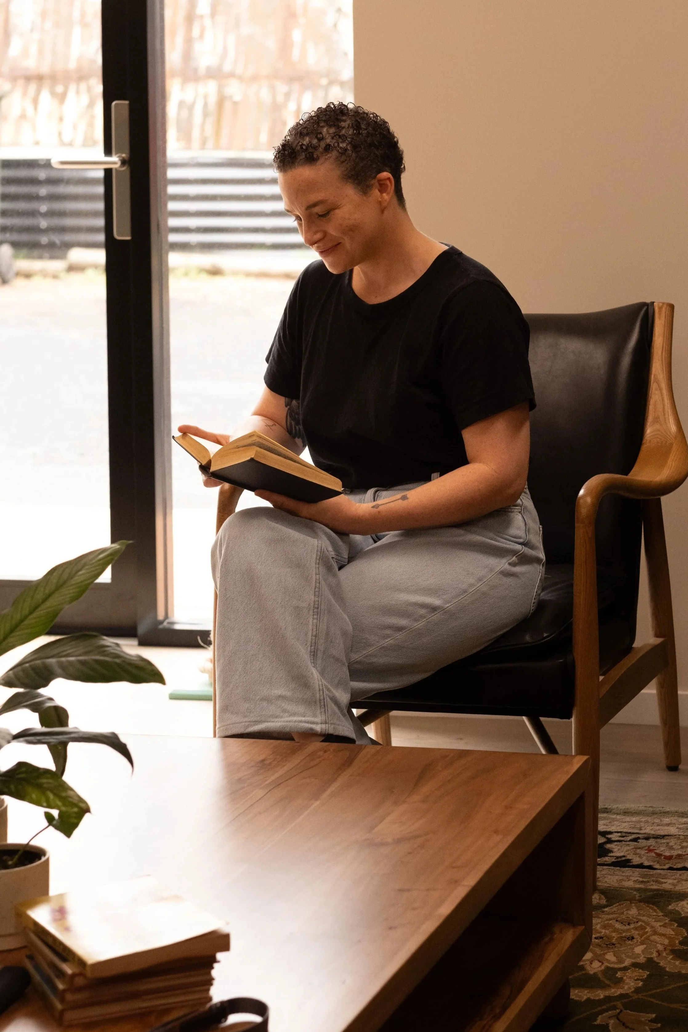 A woman with short curly hair sitting in a chair, reading a book inside a room with natural light coming through a glass door, with plants and furniture nearby.
