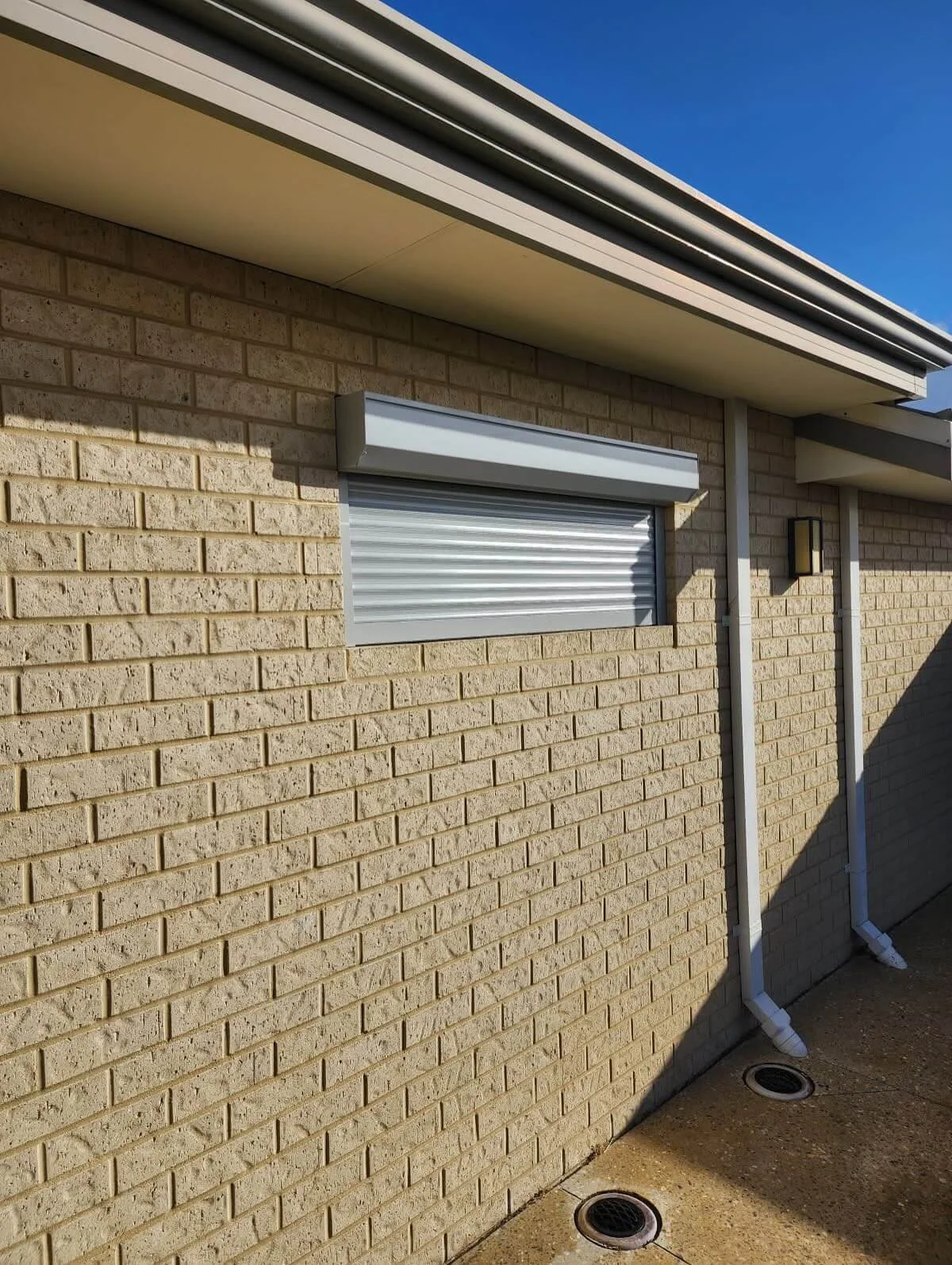 Exterior of a building with a brick wall, a small window with a metal shutter, two drainpipes, and two circular ground drains, under a clear blue sky.